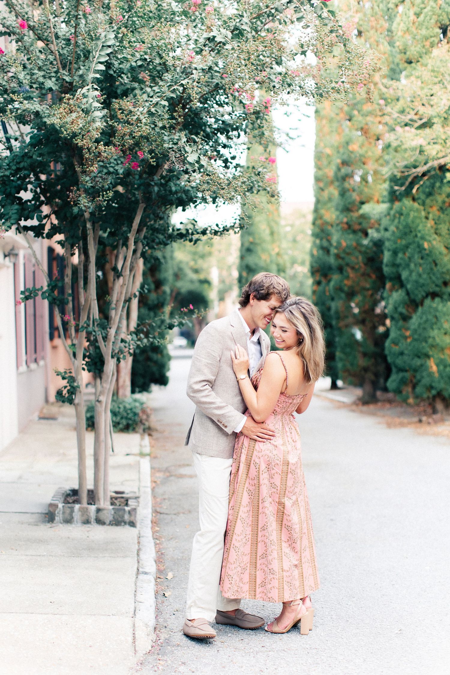 Couple embracing on a tree-lined sidewalk, woman in pink floral dress, man in tan blazer nuzzling her cheek.