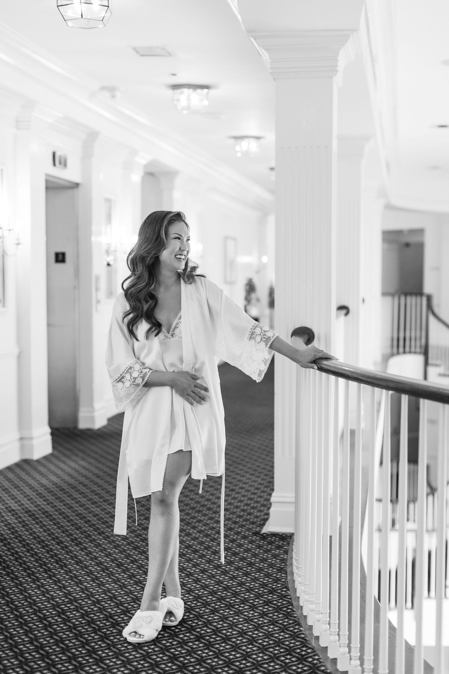 A bride in a white robe and slippers leans on a railing in an elegant hotel hallway, smiling joyfully.