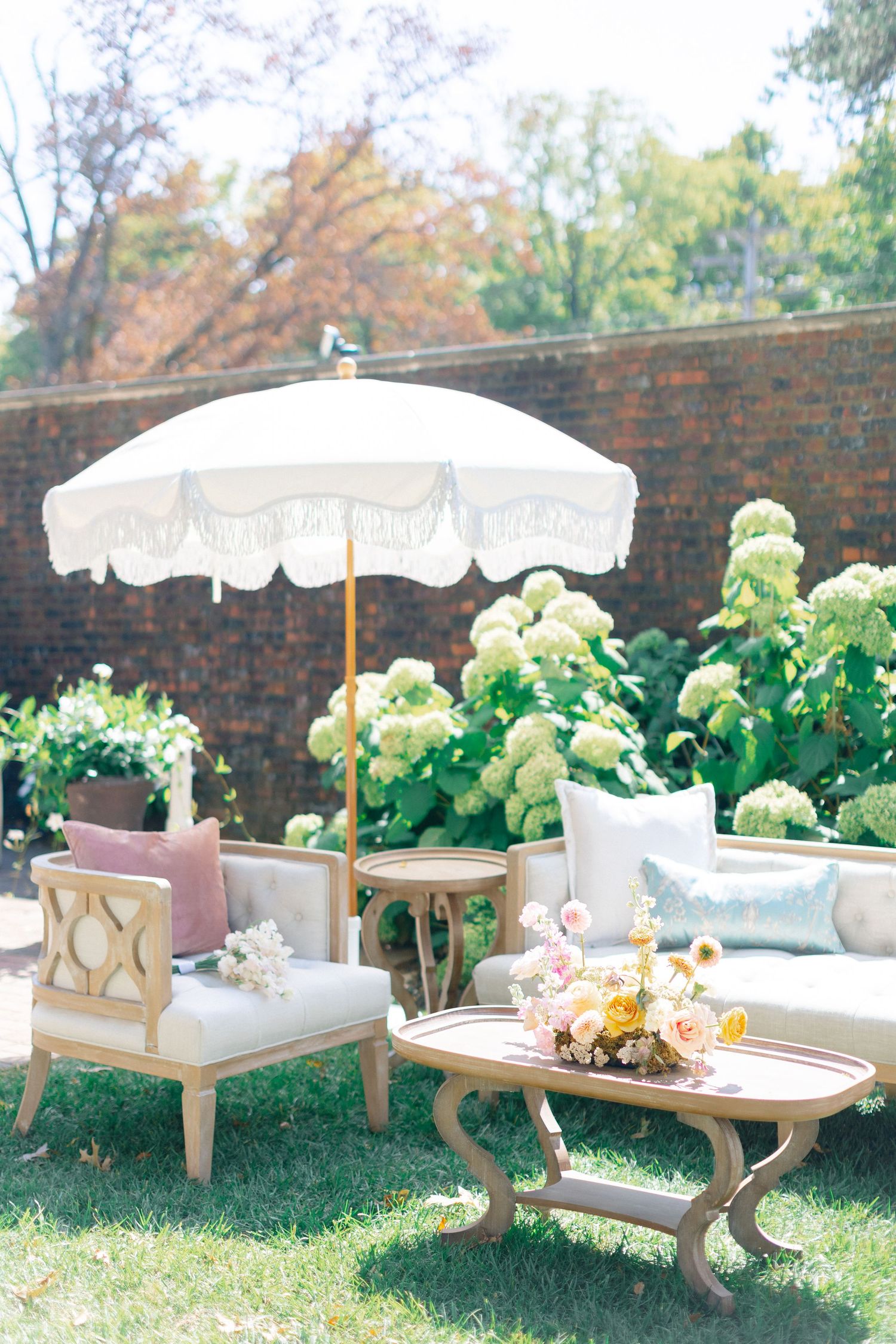 Romantic outdoor wedding lounge with white fringed umbrella, vintage furniture, pastel pillows, and hydrangea backdrop.