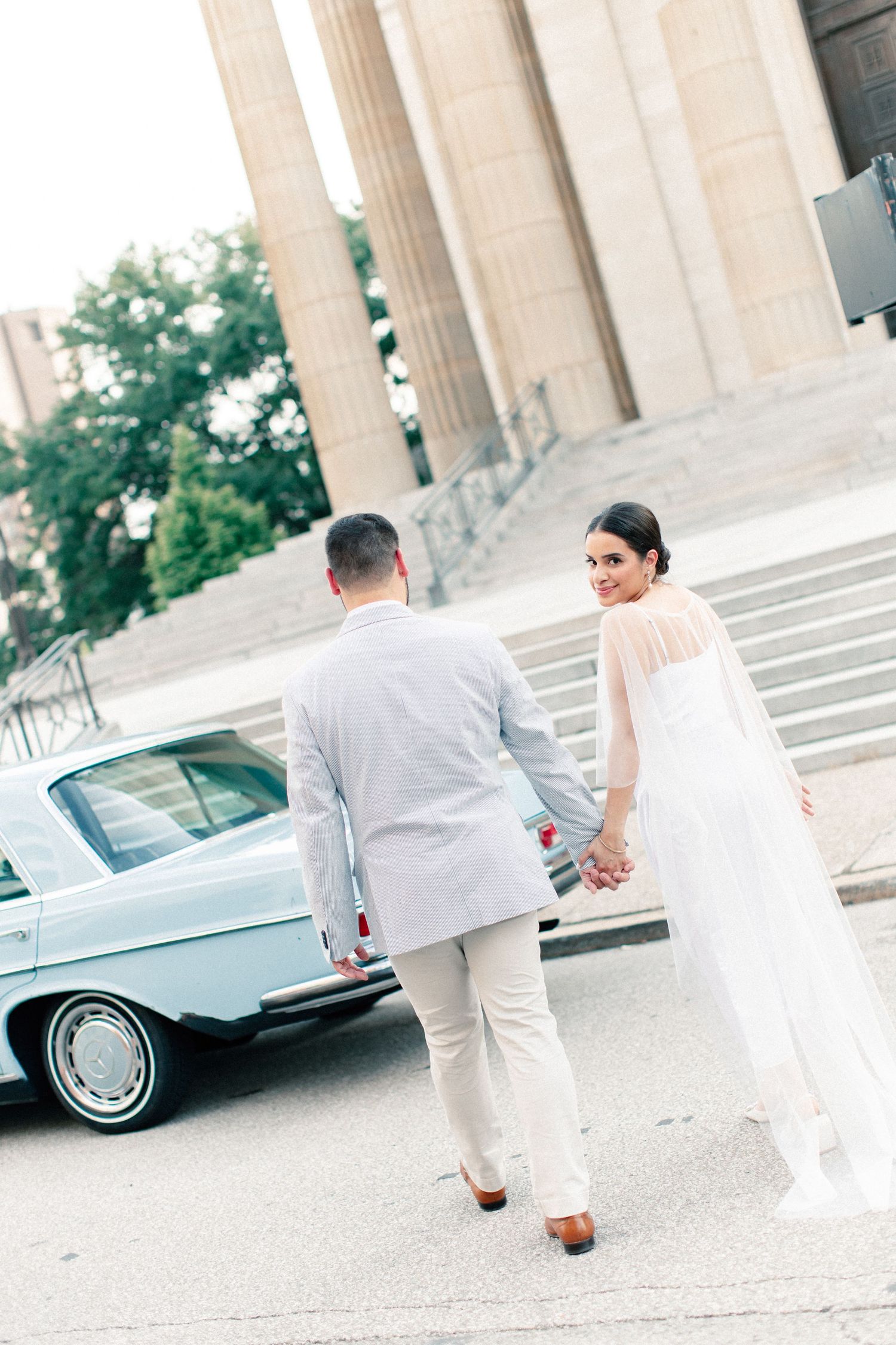 Bride and groom walk hand in hand toward a classic blue car beside grand columns at their wedding venue.