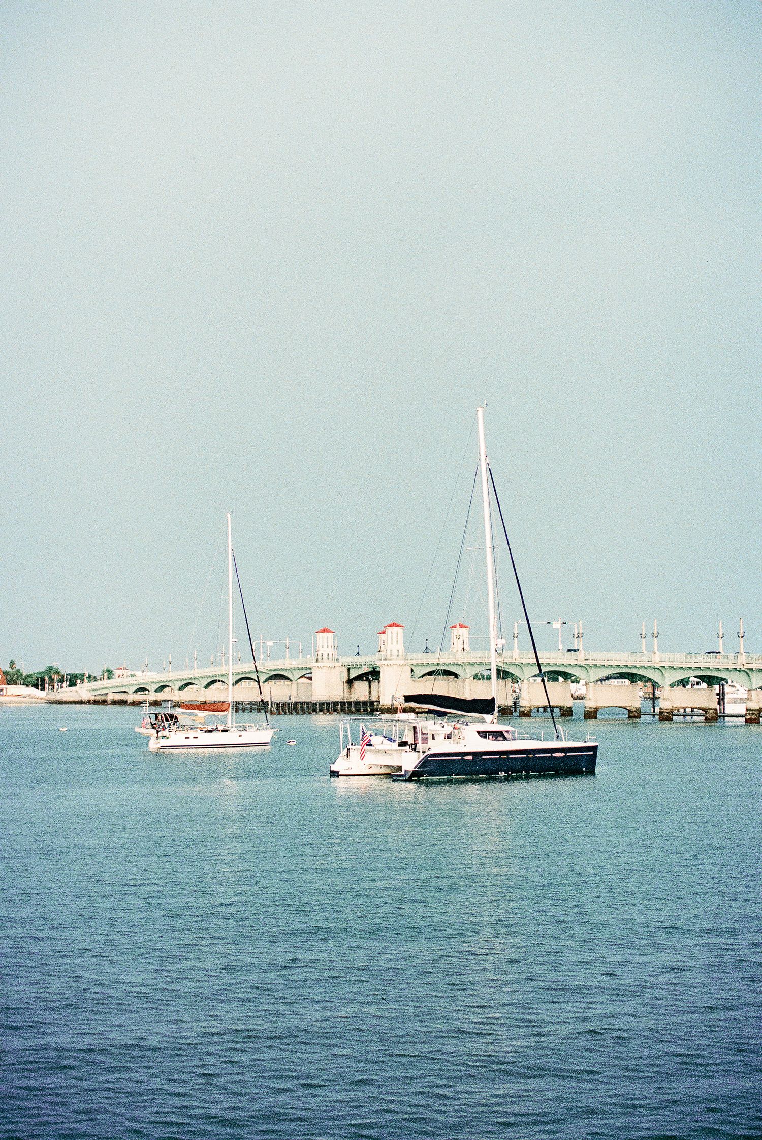 Sailboats anchored in calm blue waters near a historic arched bridge on a clear day.
