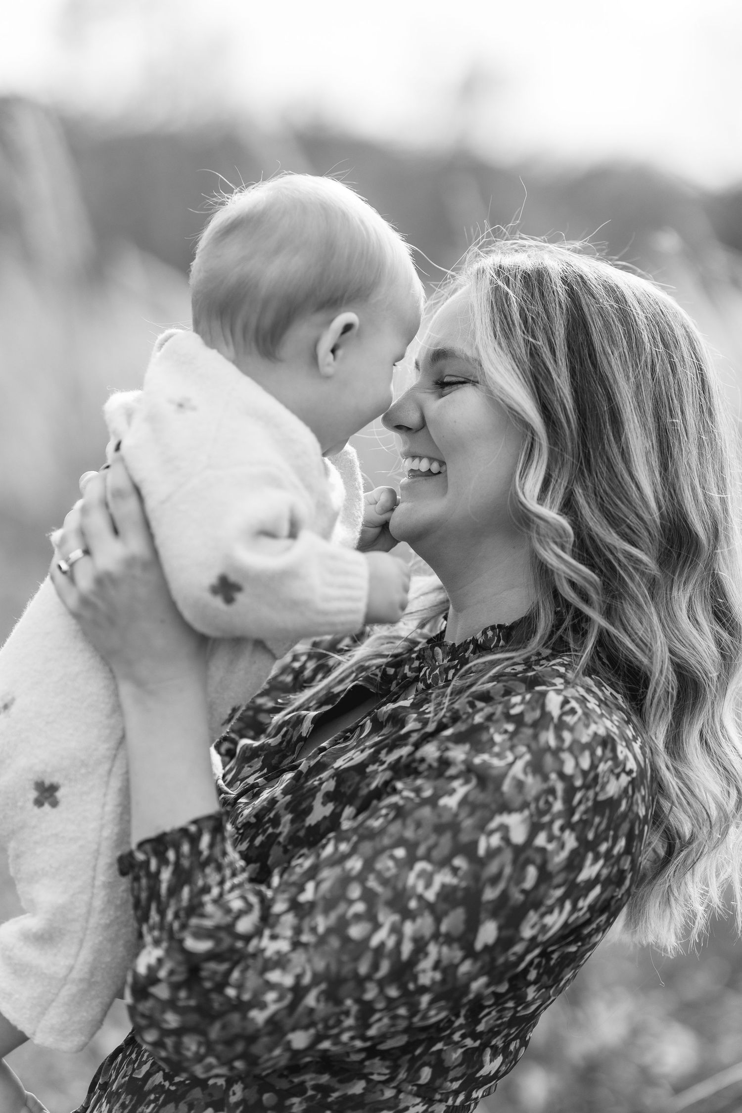 Black and white photo of a smiling woman holding a baby close, touching noses in a tender outdoor moment.
