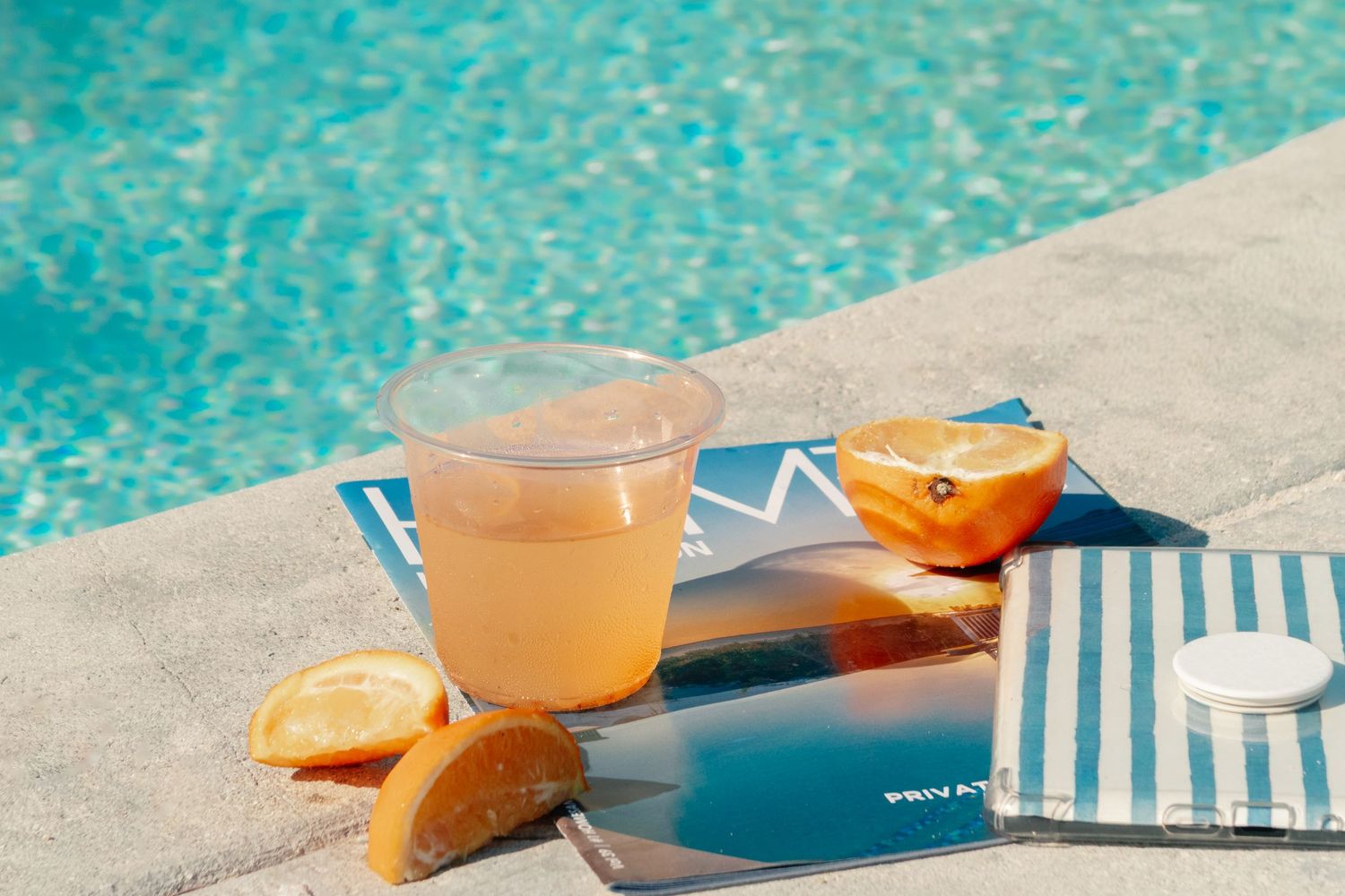 A refreshing pink drink in a plastic cup beside sliced oranges, a magazine, and striped towel by a sunny pool.