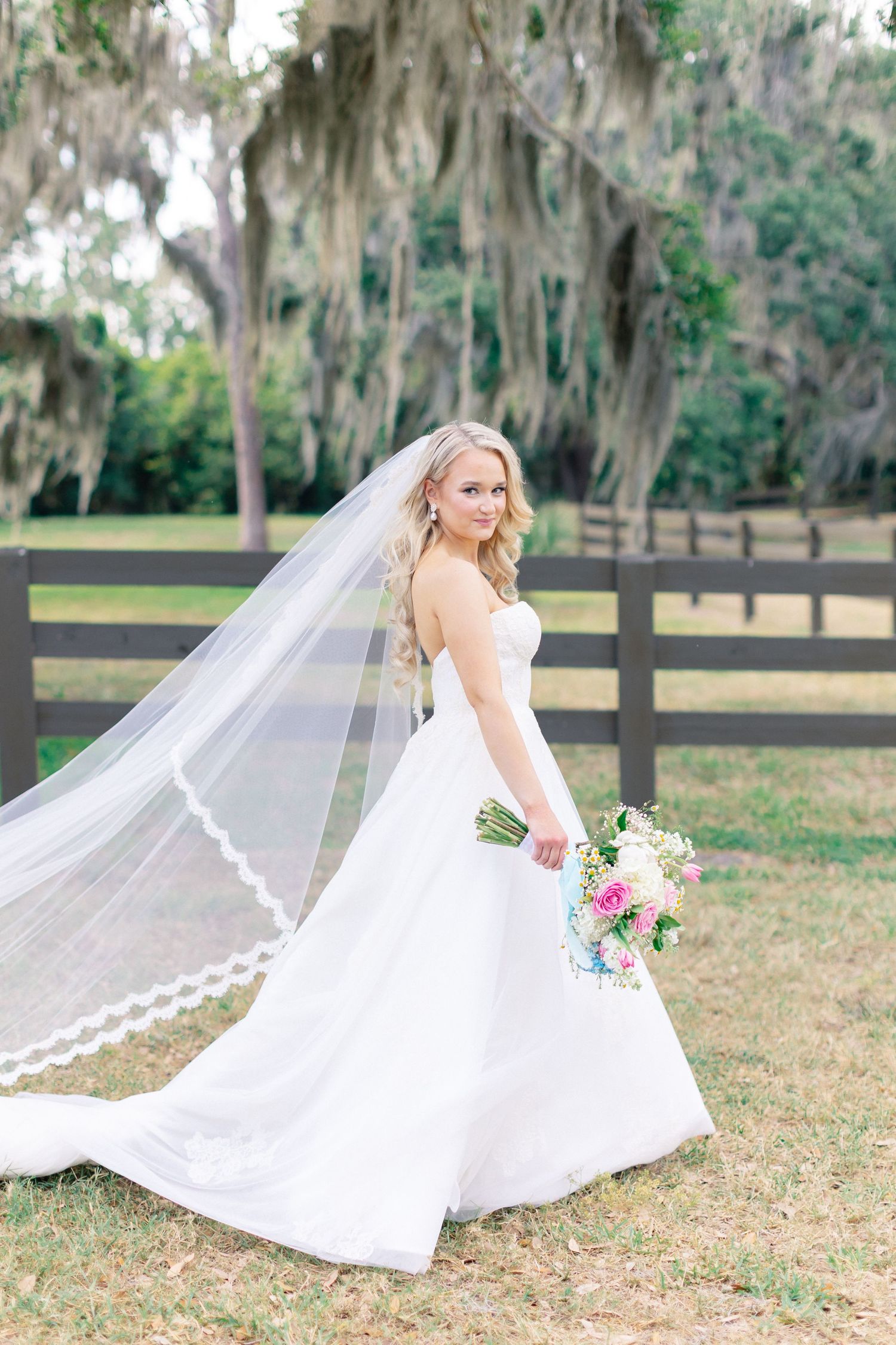 Bride in strapless white gown with lace-edged veil holding pink bouquet, posing outdoors near wooden fence and oak trees, Bella Cosa