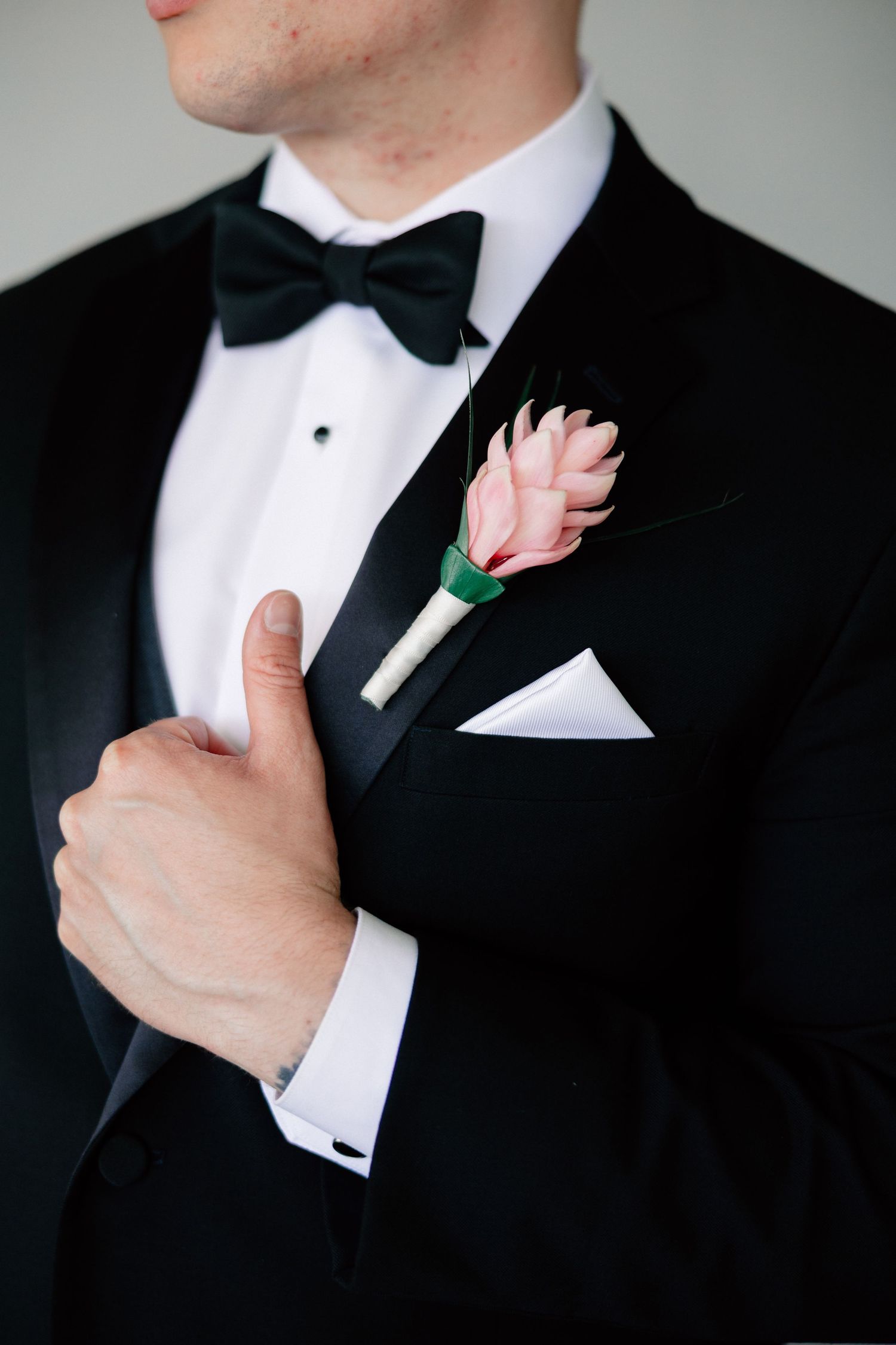 Groom in black tuxedo with bow tie, pink boutonniere, and white pocket square, holding lapel elegantly.