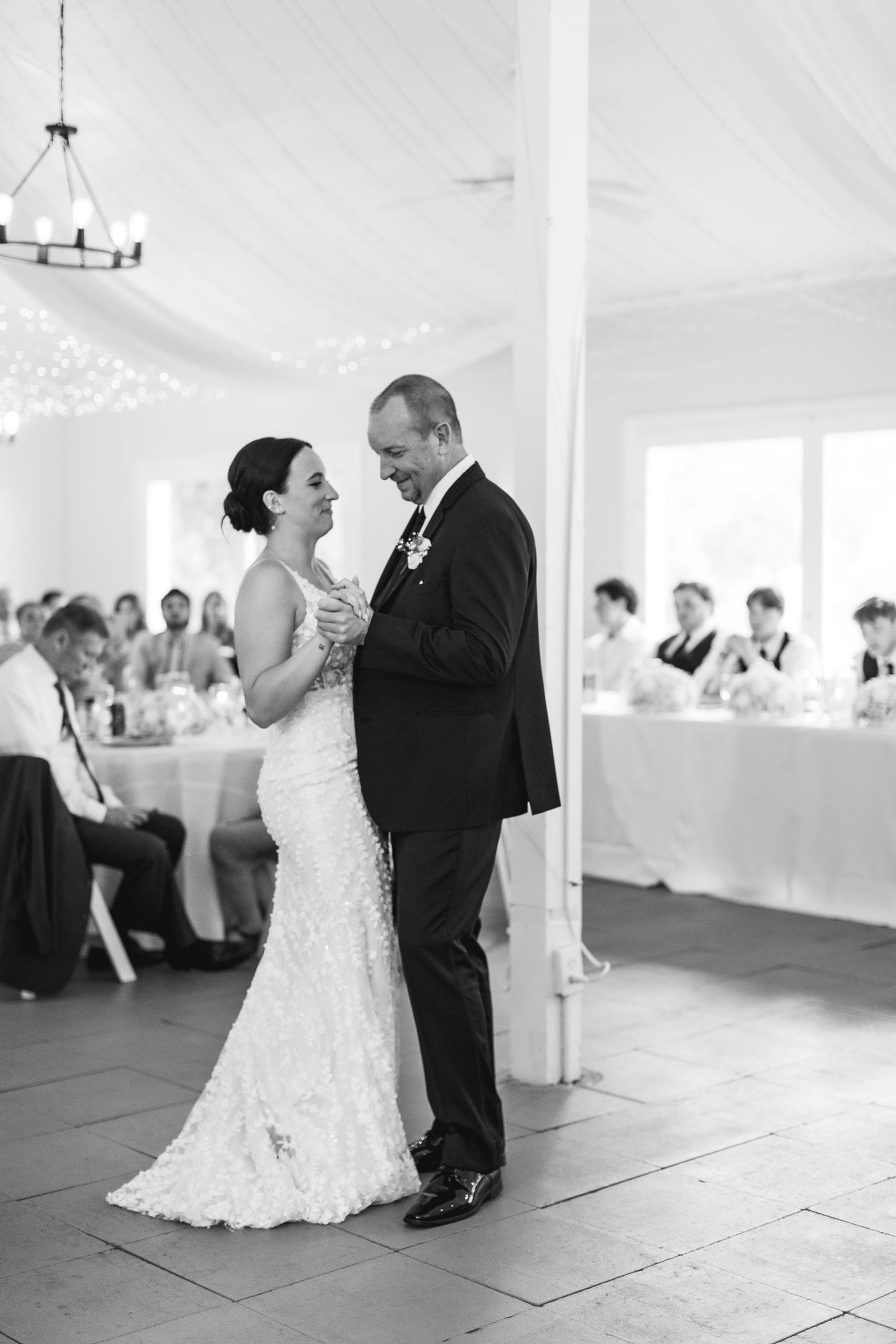 Black and white photo of bride and groom sharing first dance at wedding reception with guests seated in background.