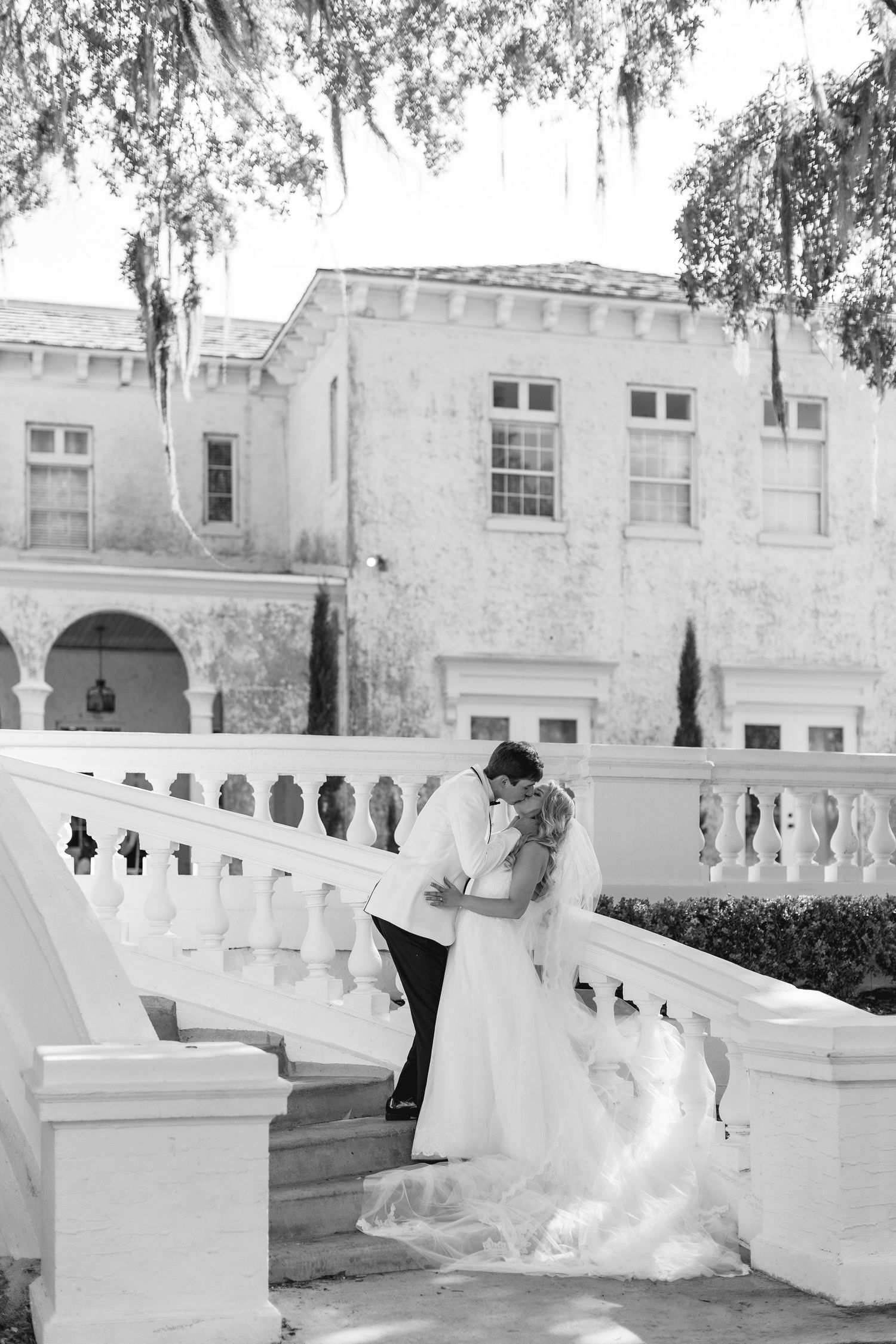 Black and white photo of a bride and groom sharing a kiss on elegant white staircase steps outside a grand estate, wedding at Bella Cosa