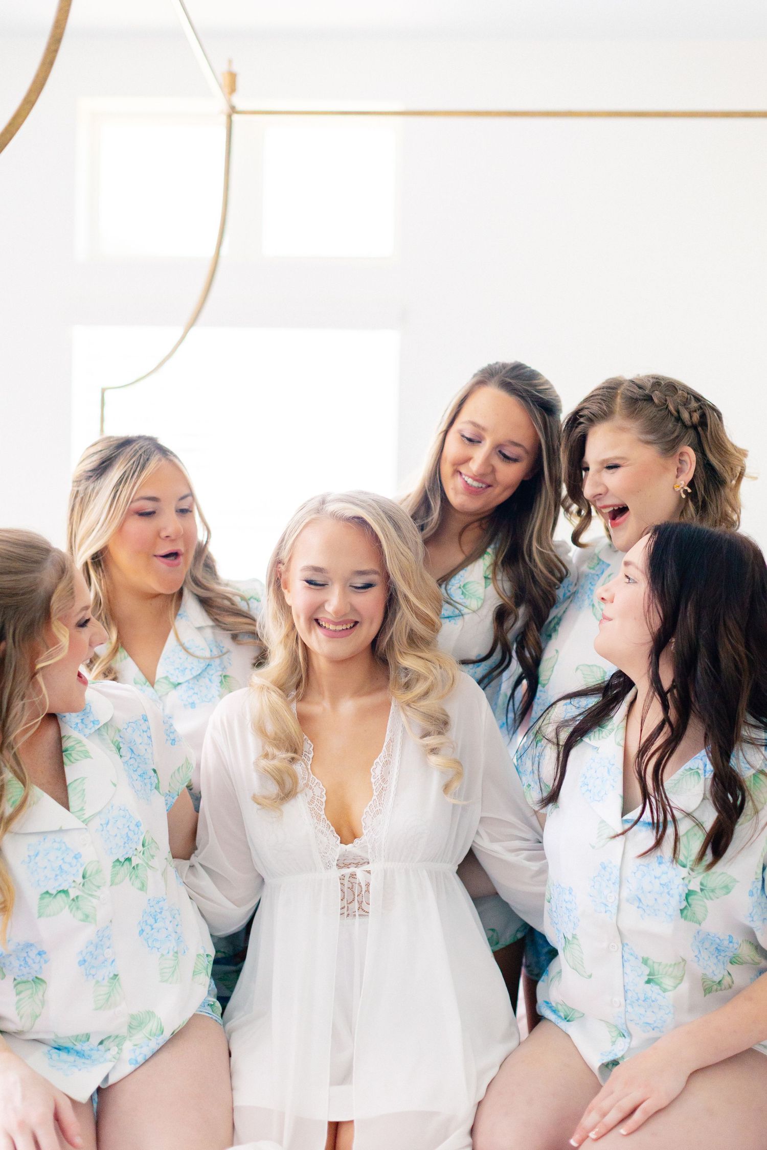 Bride in white lace robe surrounded by bridesmaids in matching floral pajamas laughing together before wedding, wedding at Bella Cosa