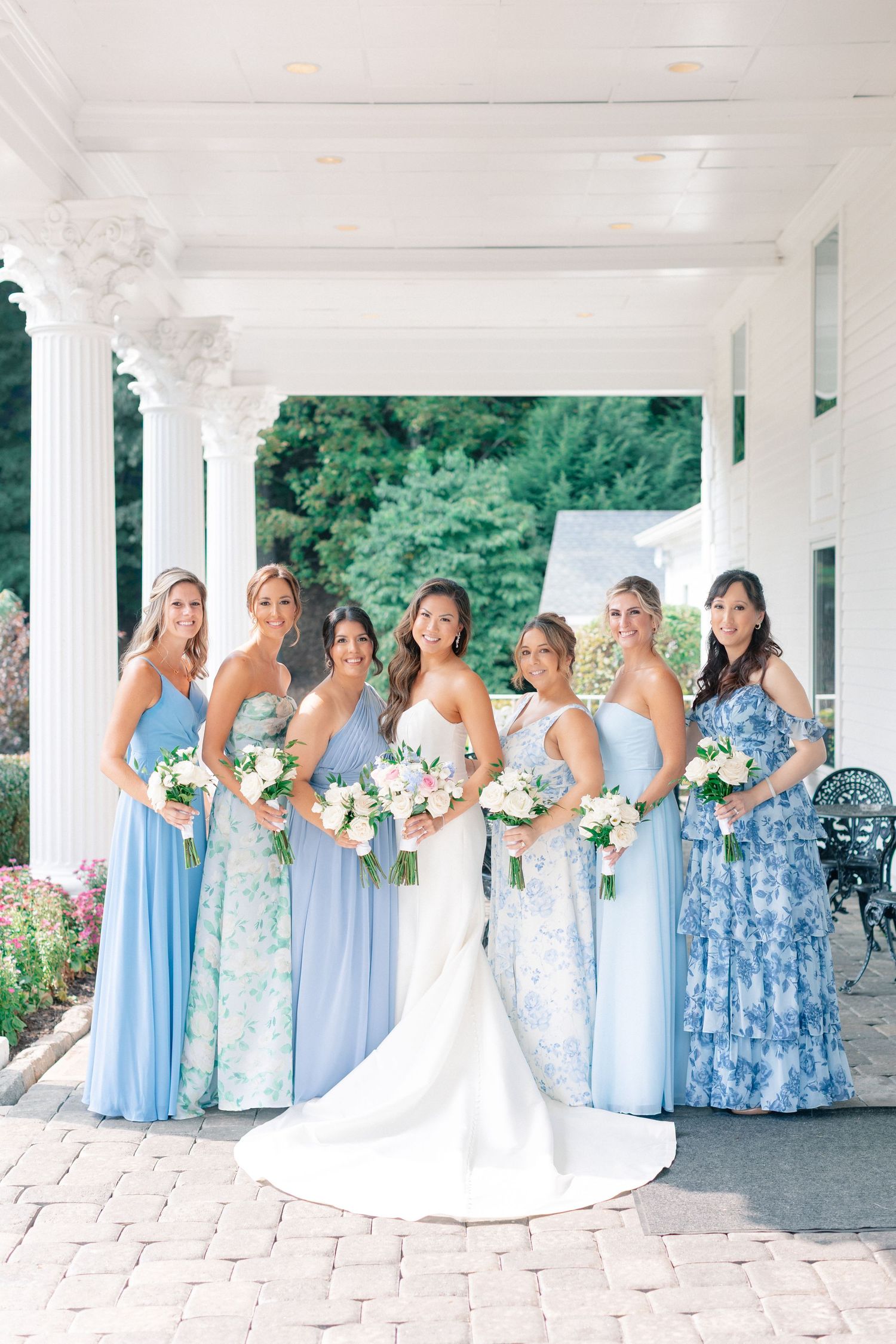 Bride and bridesmaids in blue dresses holding floral bouquets pose on a white columned porch.