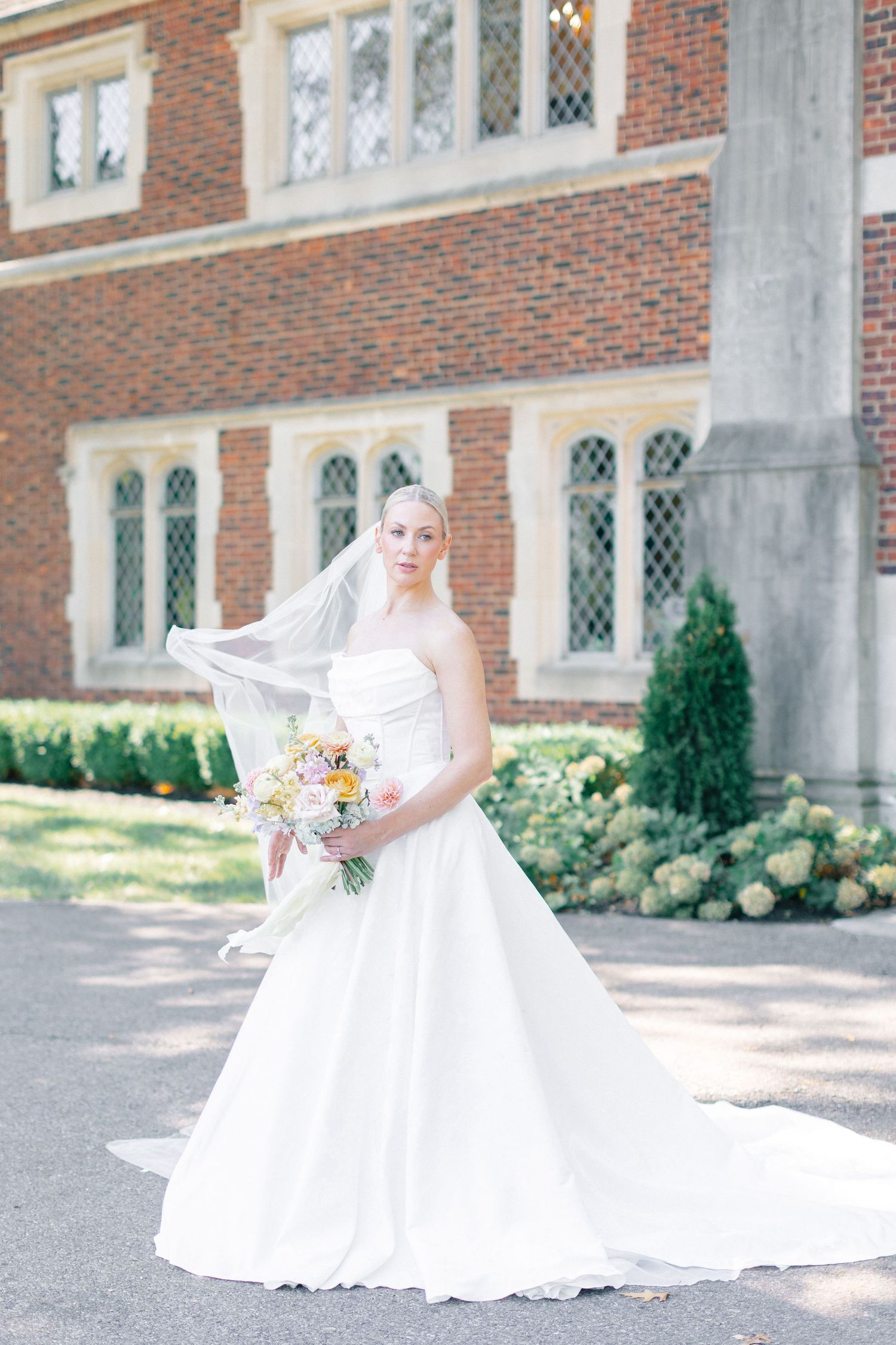 Elegant bride in strapless ball gown and veil holds colorful bouquet outside a brick manor house.