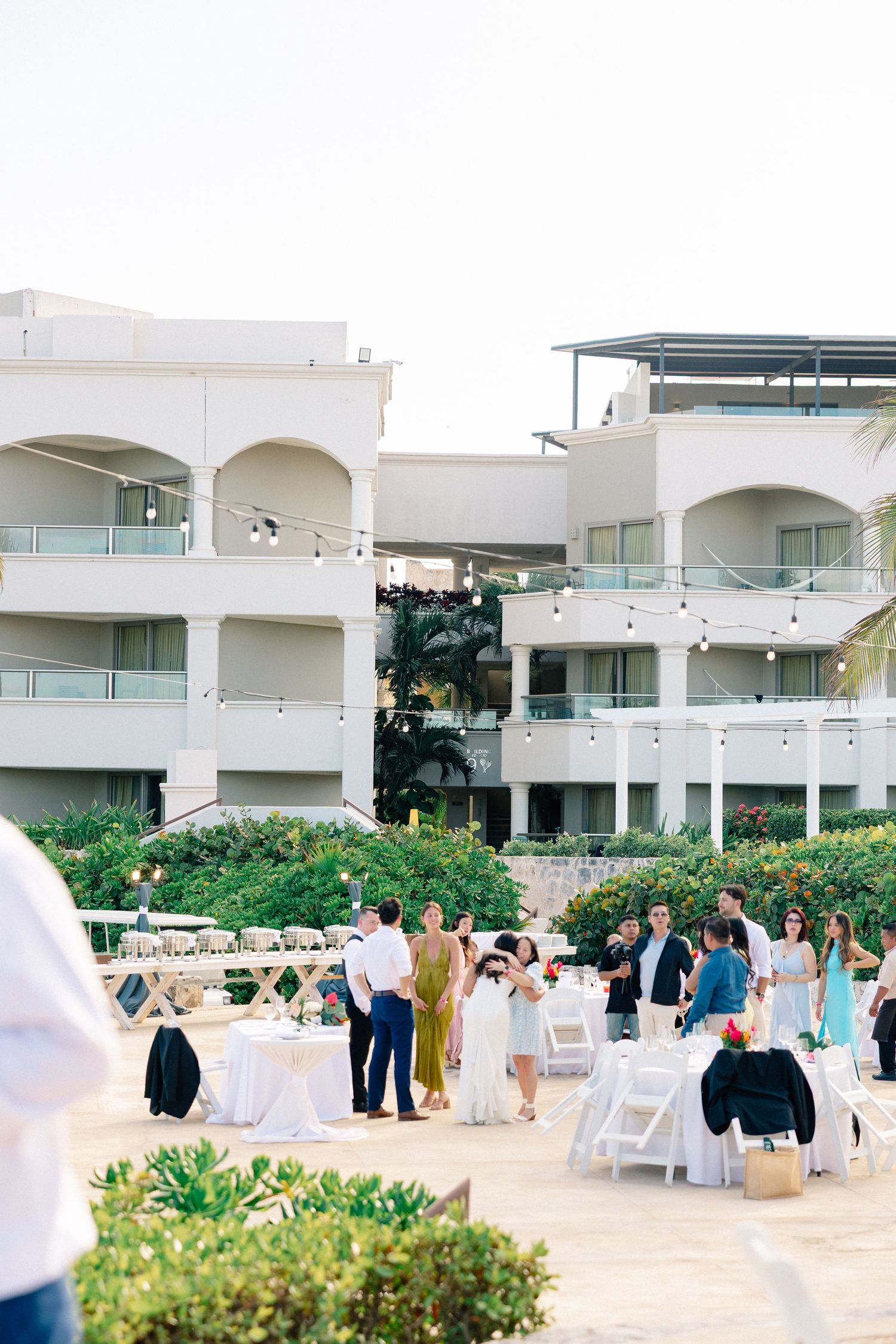 Guests mingle at an outdoor wedding reception at a white resort with arched balconies, string lights, and lush greenery.