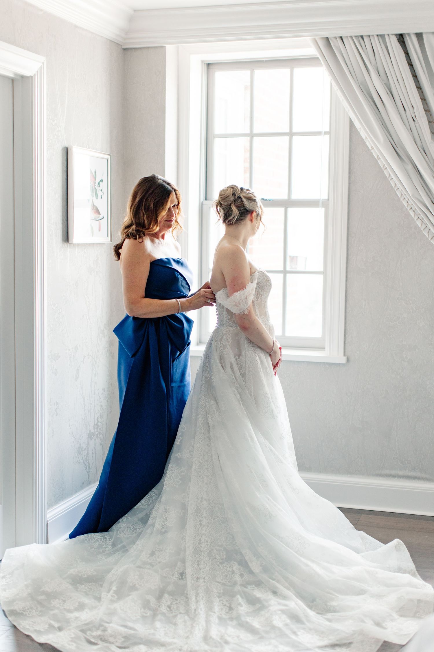 Bride in lace gown being helped by bridesmaid in blue strapless dress near bright window on wedding day.
