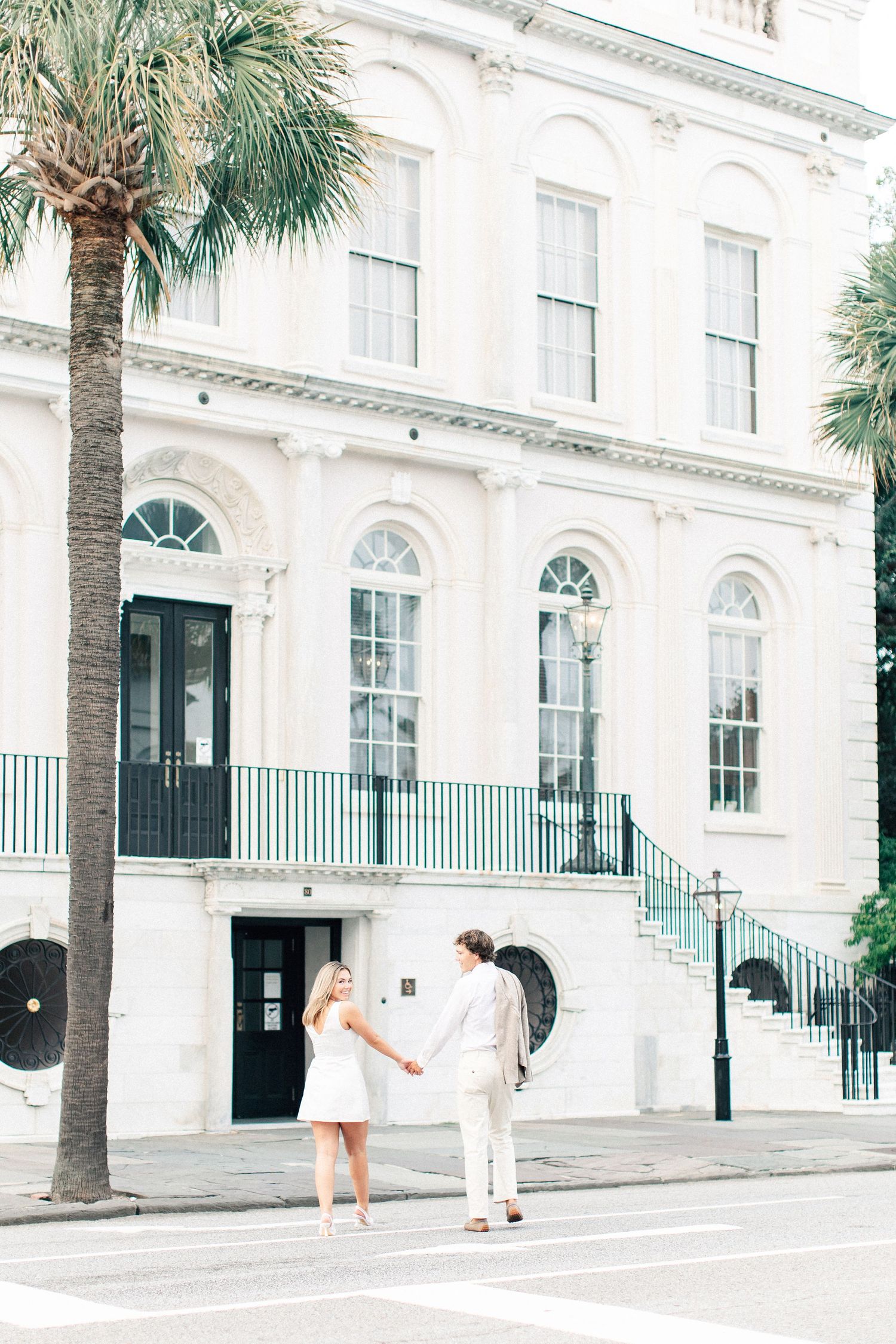 Couple holding hands in front of a grand white historic building with palm tree in Charleston, SC.