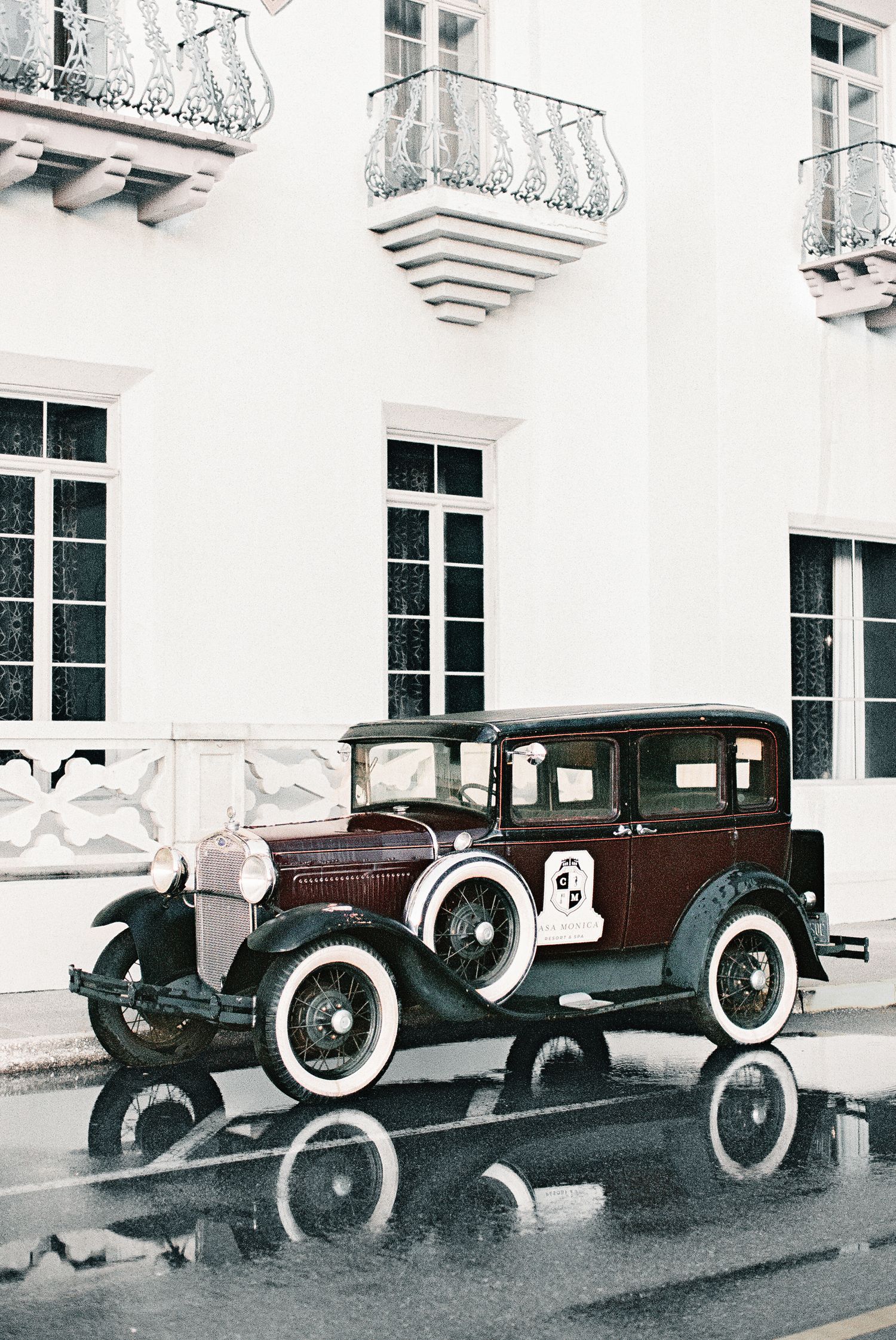 Vintage antique car parked on wet street beside white classical building with ornate balconies.