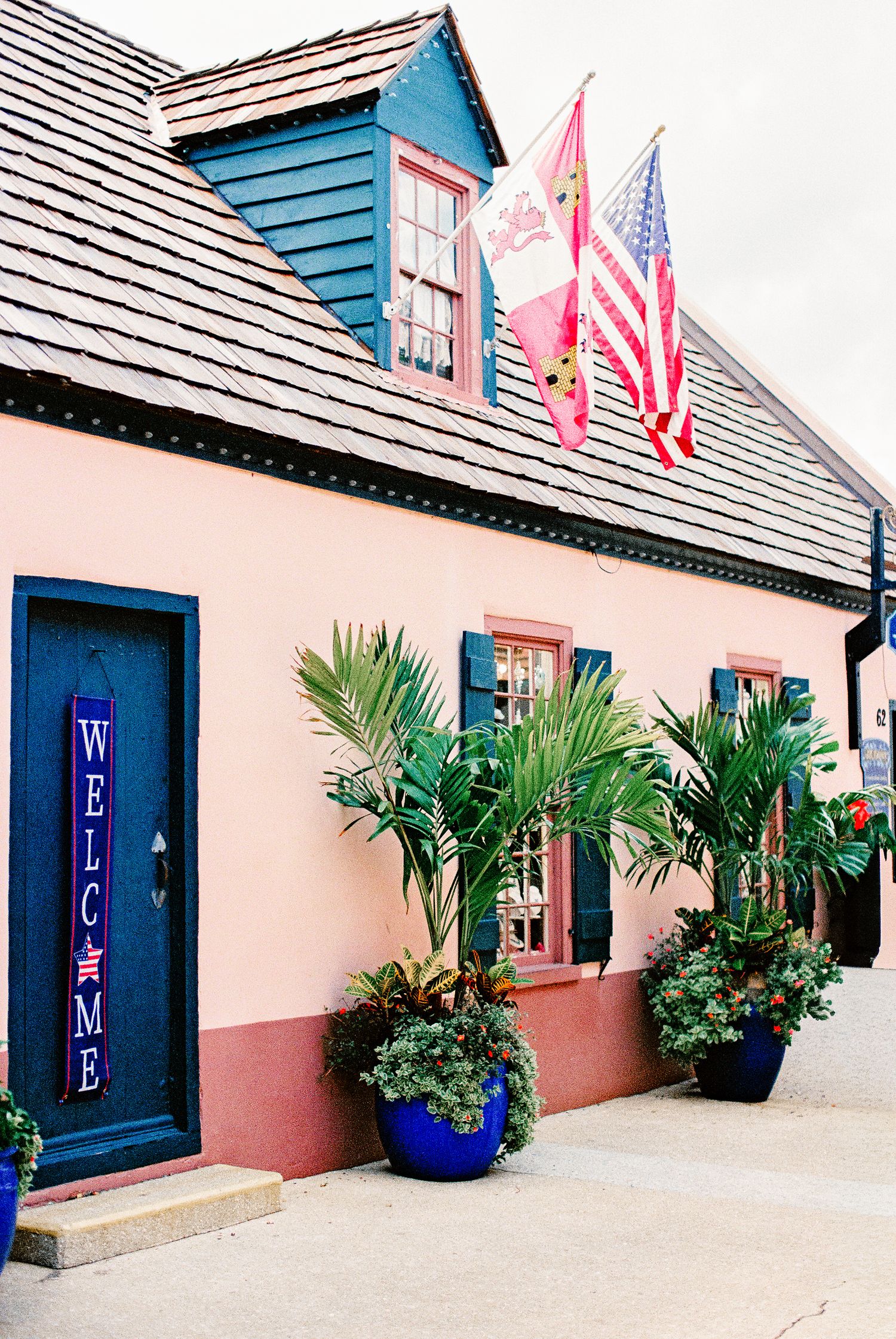 Pink building with blue door, Welcome sign, American flag, and tropical plants in blue pots outside a charming shop.