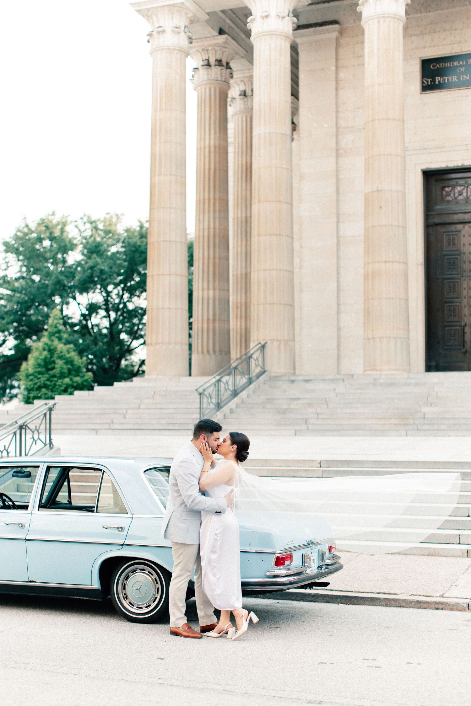Wedding couple sharing a kiss beside a vintage blue Mercedes outside a grand columned church.
