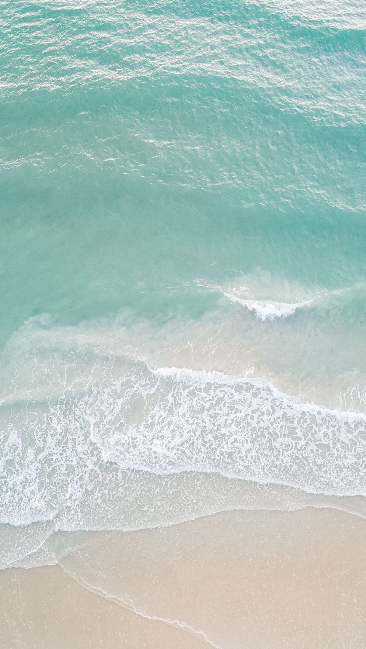 Aerial view of turquoise ocean waves gently washing onto a white sandy beach.