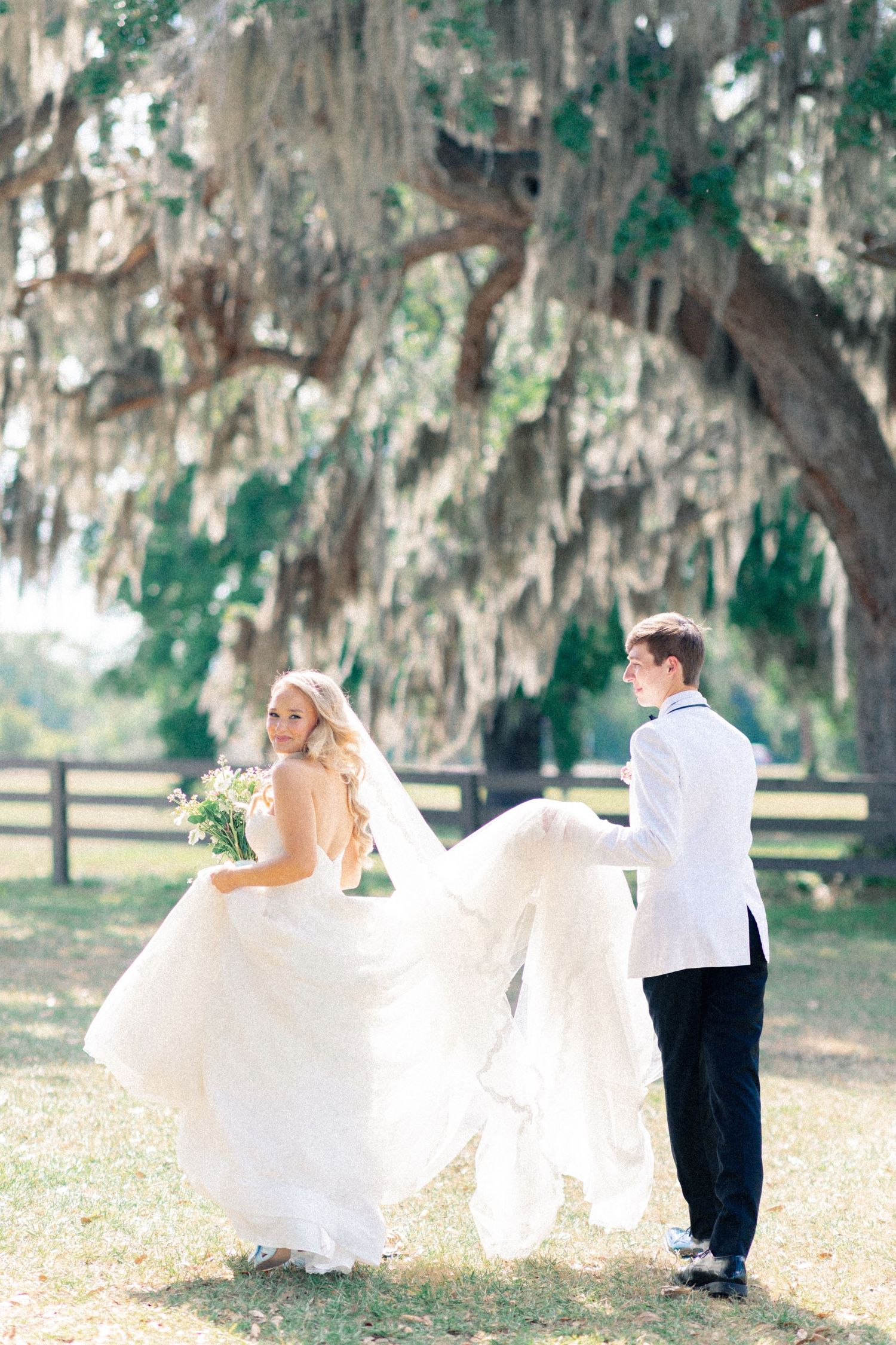 Bride and groom walking outdoors under Spanish moss-draped oak trees, groom carrying bride's flowing white wedding gown train.