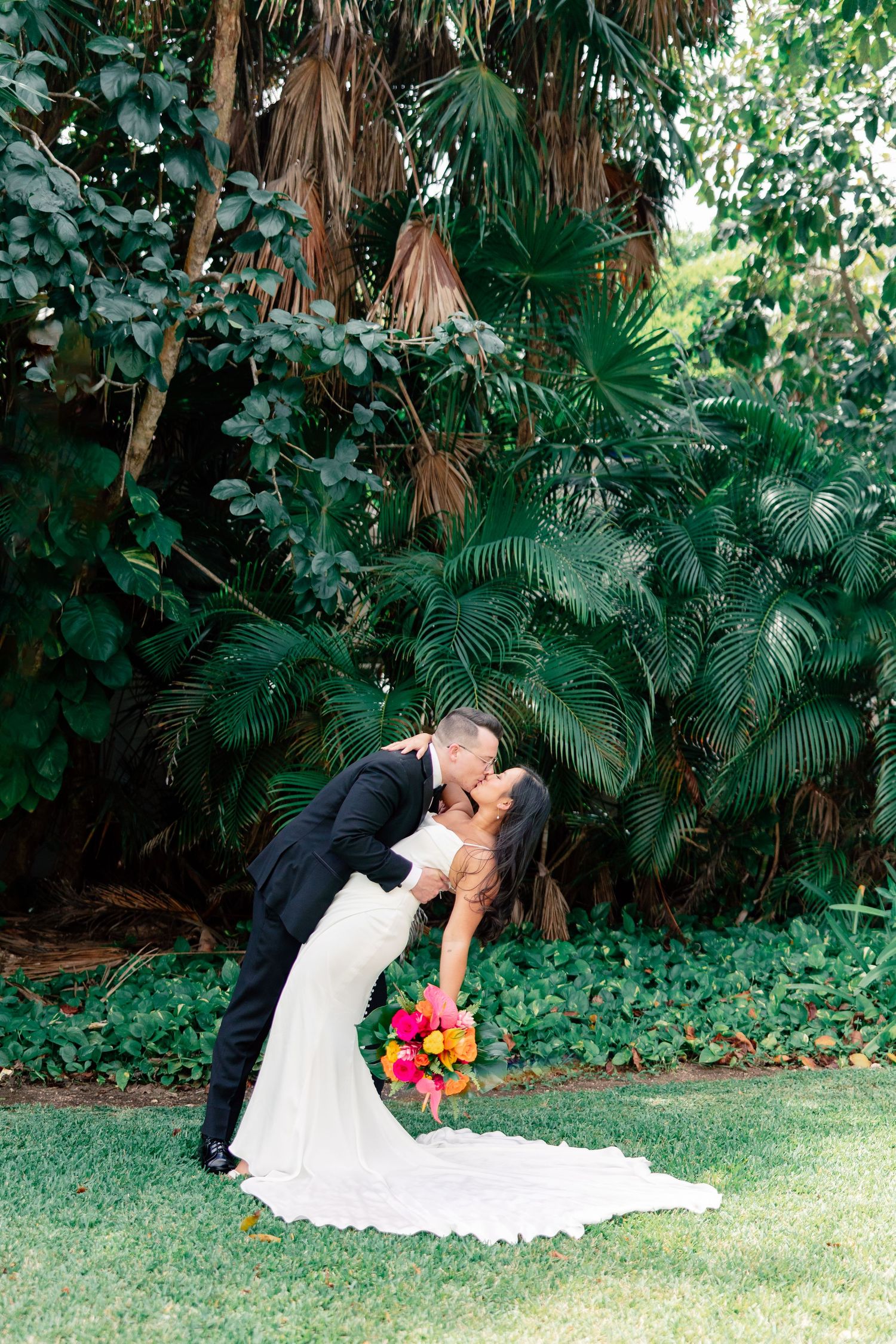 Bride and groom share a romantic dip kiss in front of lush tropical greenery, bride holding vibrant colorful bouquet.