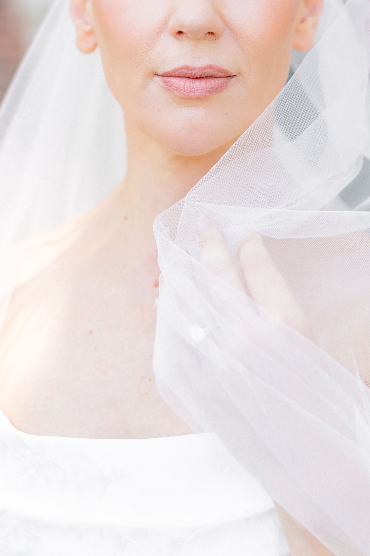 Close-up of a bride wearing a white lace dress and flowing veil, holding it delicately near her face.