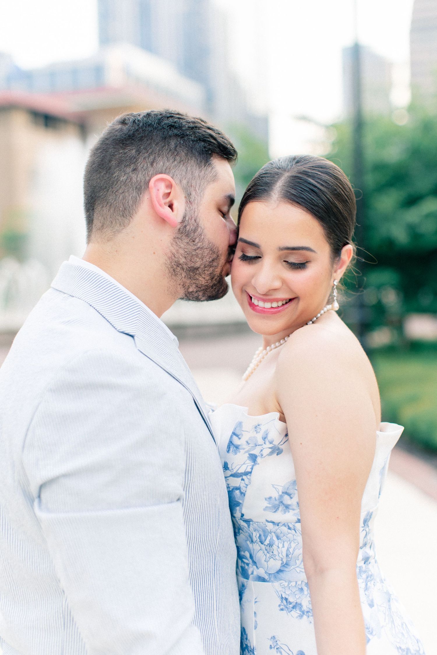 Groom tenderly kissing bride on the cheek as she smiles in a blue floral dress with city backdrop.