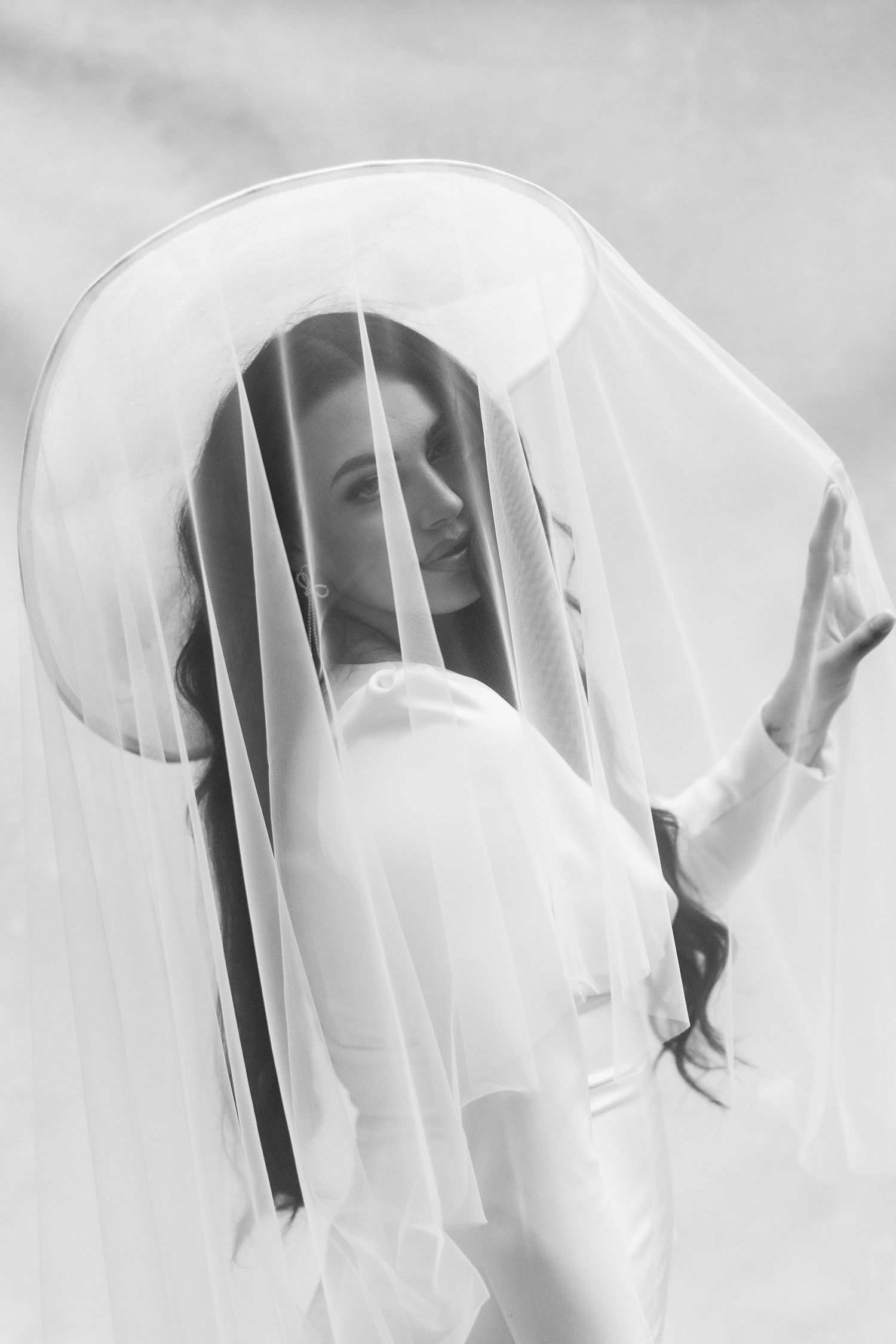 Black and white portrait of bride wearing wide-brim hat with flowing veil draped over face.