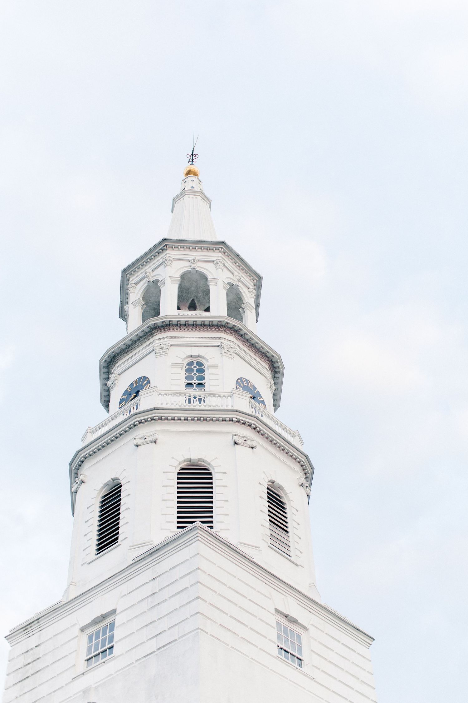 White colonial church steeple with ornate architectural details and gold finial against a pale blue sky.