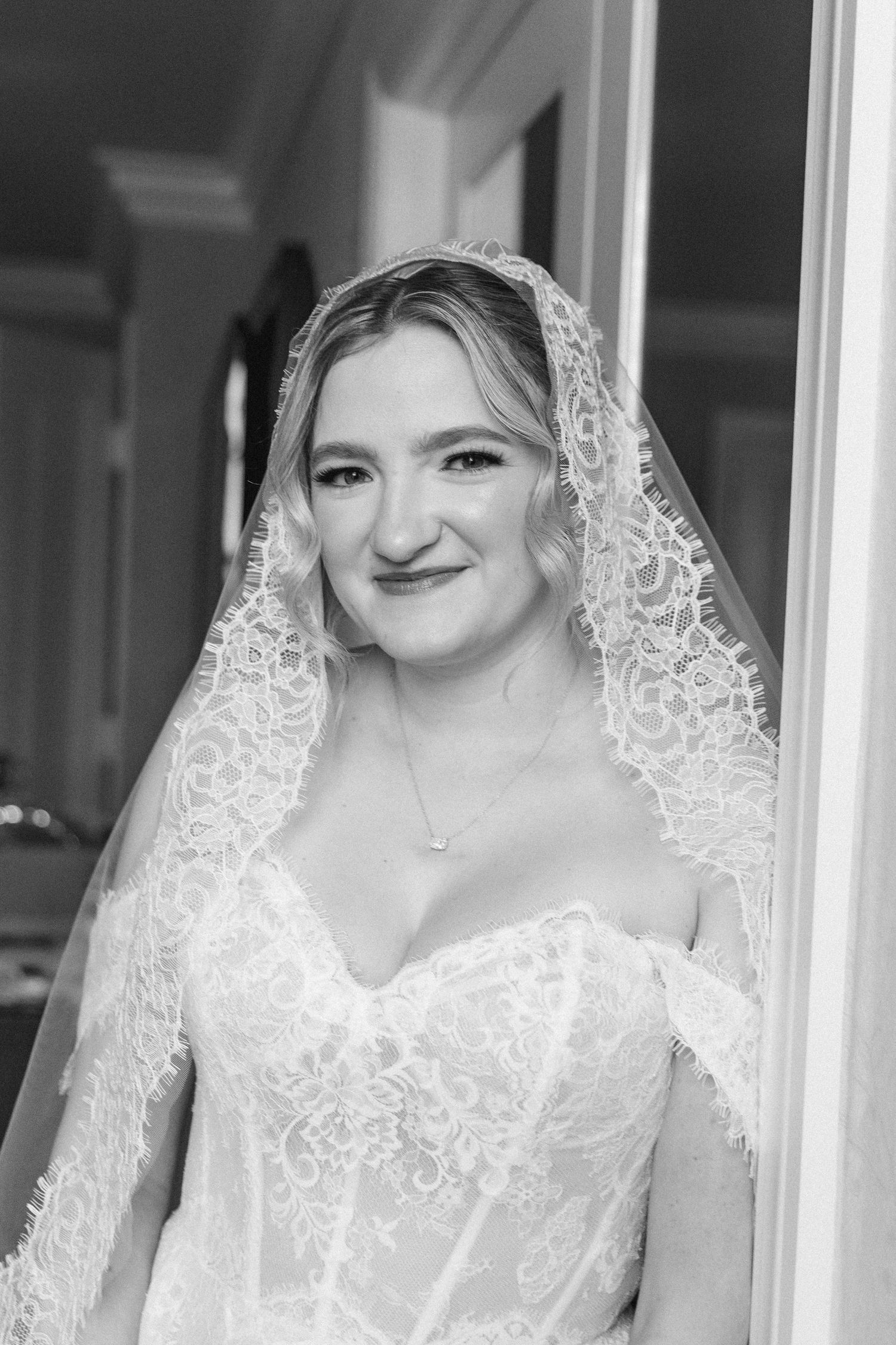 Black and white portrait of a bride wearing a lace veil and strapless wedding gown, smiling softly indoors.