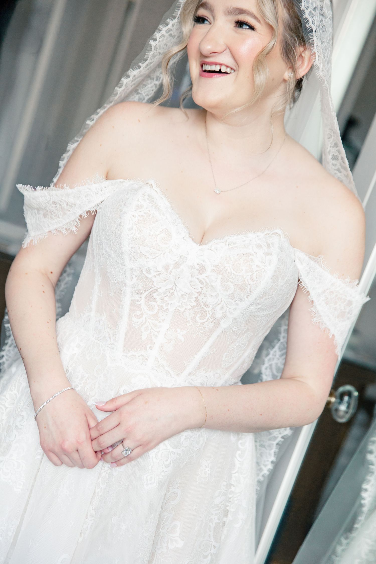Smiling bride in off-shoulder lace wedding gown with veil, showcasing elegant bridal style.