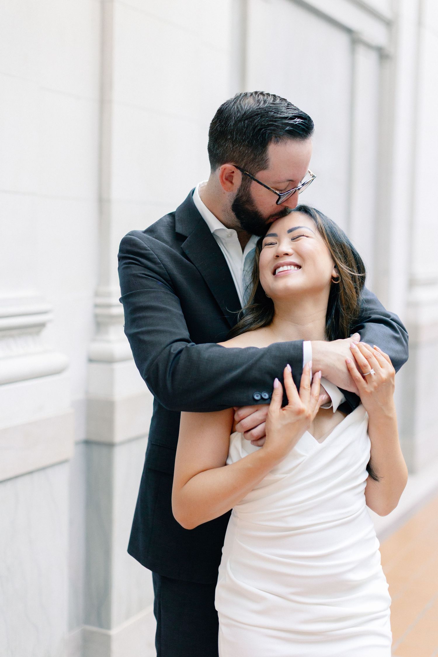 A groom in a dark suit embraces his smiling bride in a white dress, sharing a tender kiss on her head.