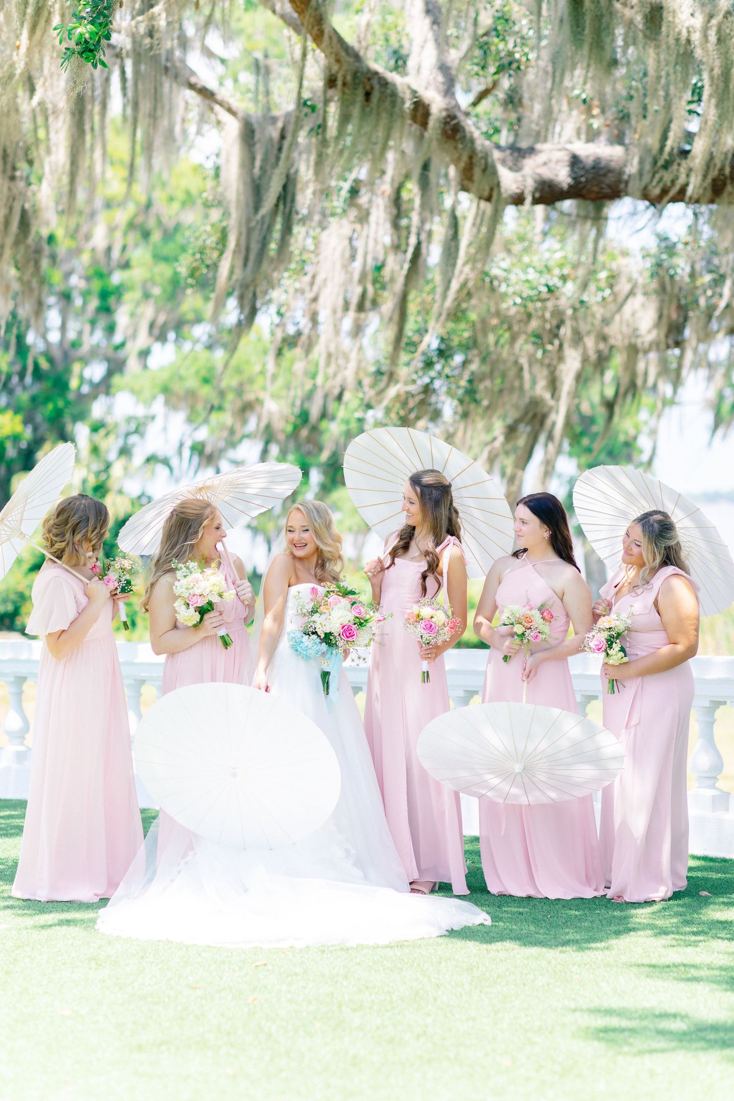 Bride in white gown with bridesmaids in pink dresses holding parasols and floral bouquets under moss-draped oak trees, wedding at Bella Cosa