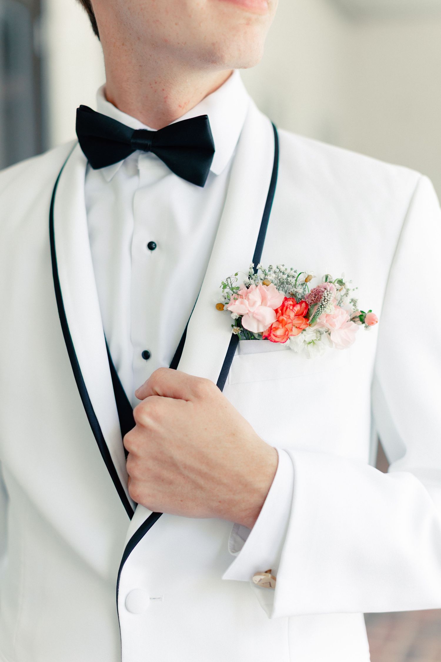 Groom in white tuxedo with black bow tie and floral boutonniere featuring pink and coral flowers, wedding at Bella Cosa