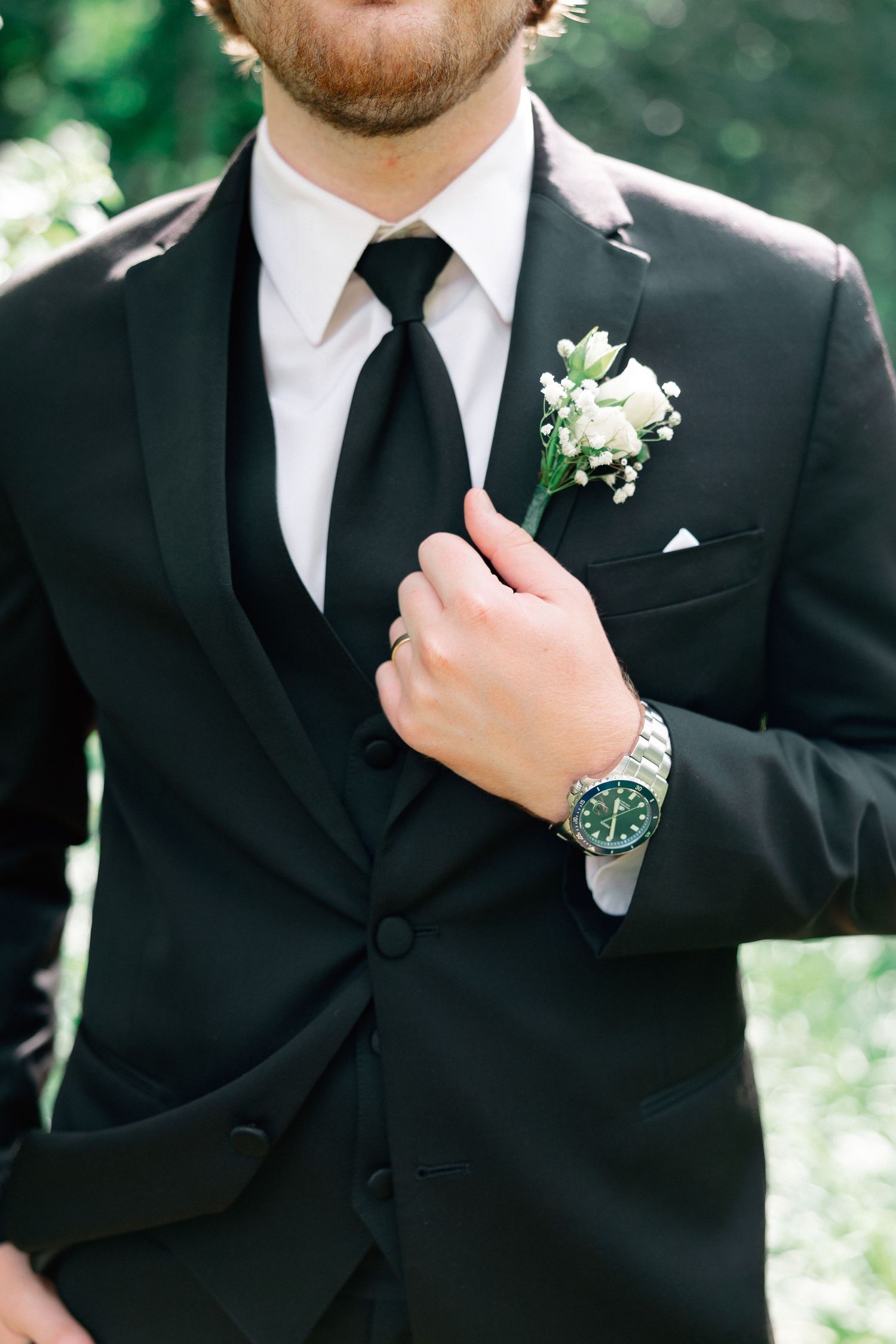 Groom in black tuxedo with white boutonniere and silver watch adjusting his tie outdoors.