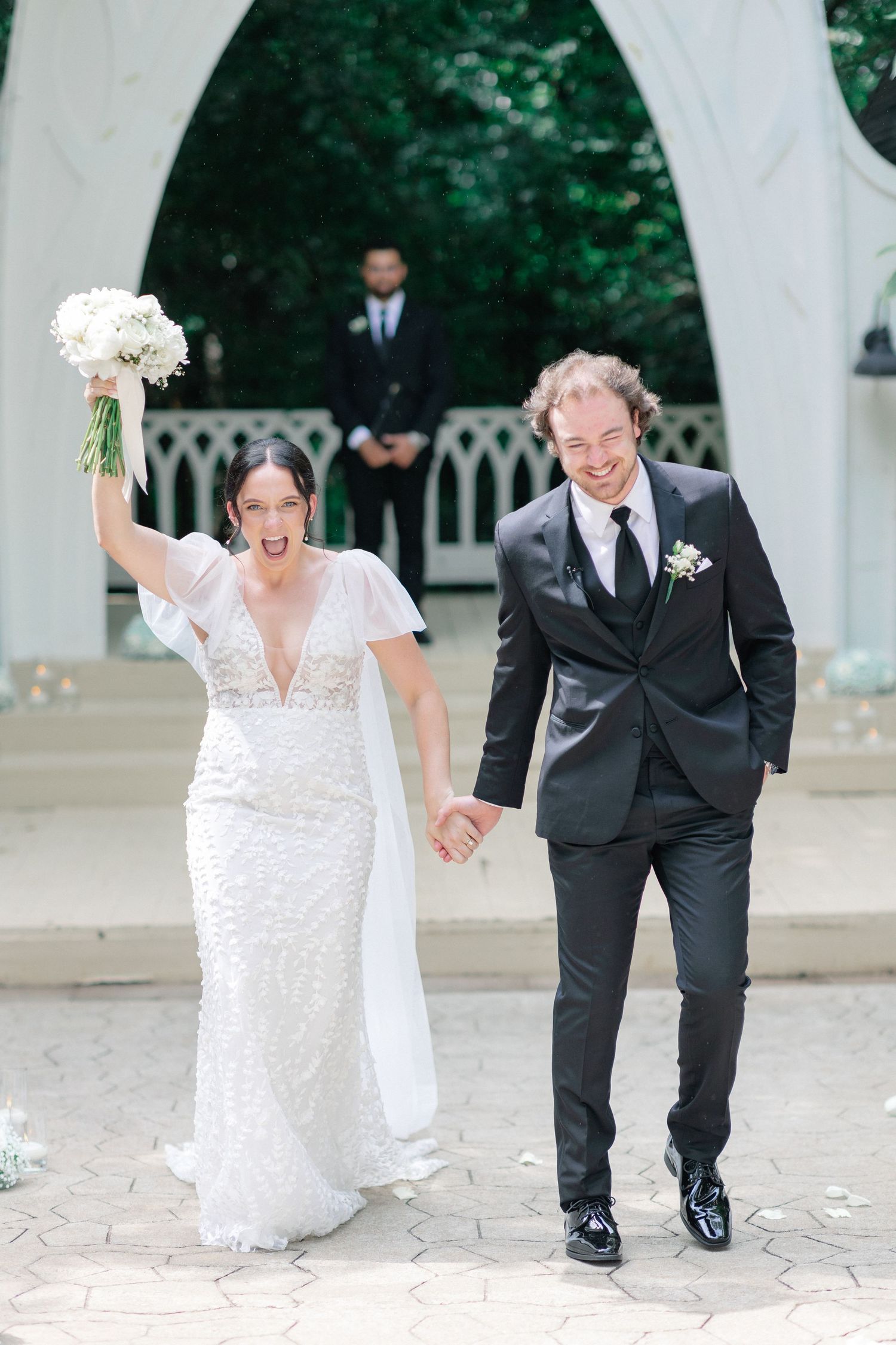 Joyful bride in white lace gown raises bouquet while groom in black tuxedo smiles, celebrating after outdoor wedding ceremony.