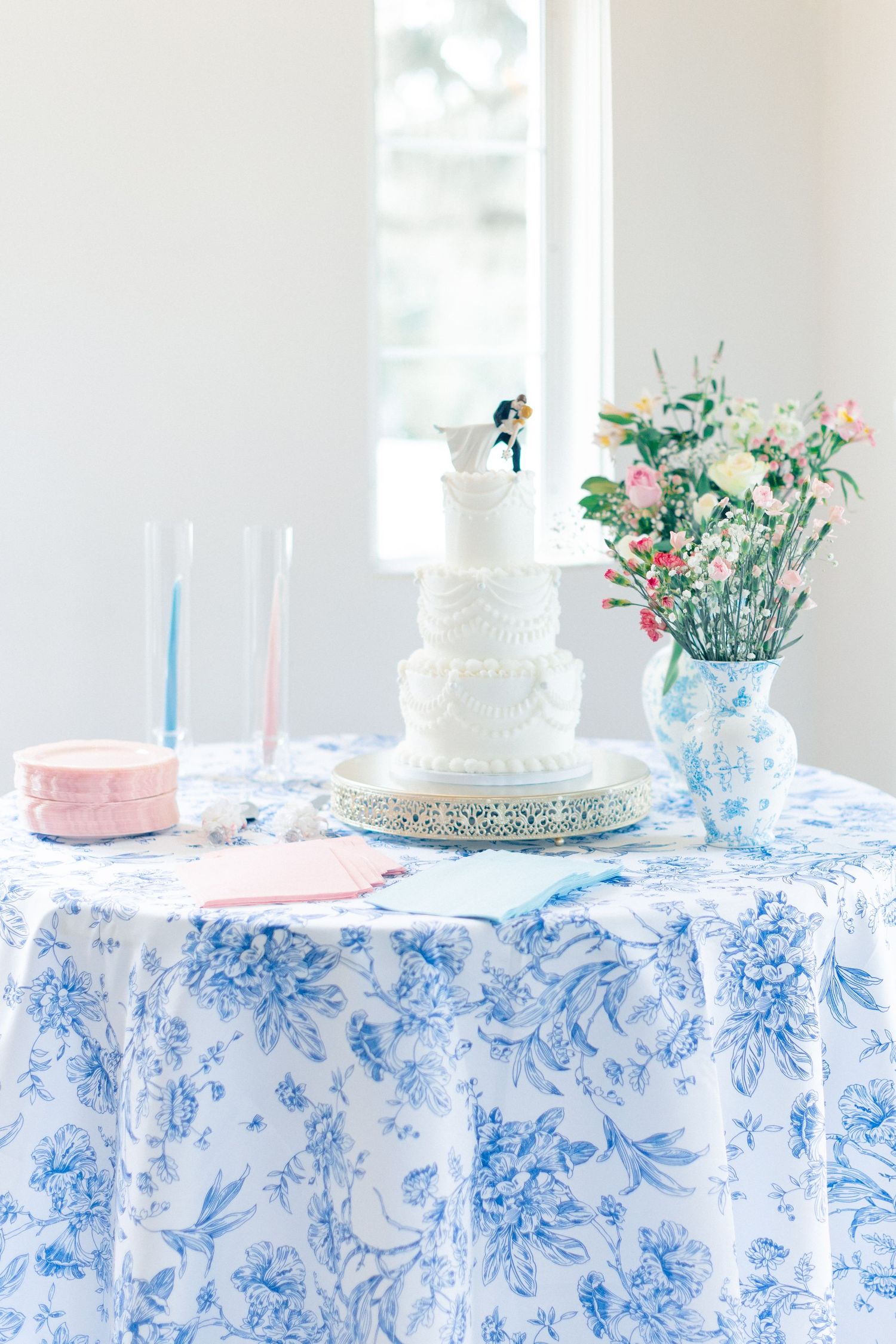 White tiered wedding cake with bride and groom topper on blue floral tablecloth with pink roses in vintage pitcher, wedding at Bella Cosa