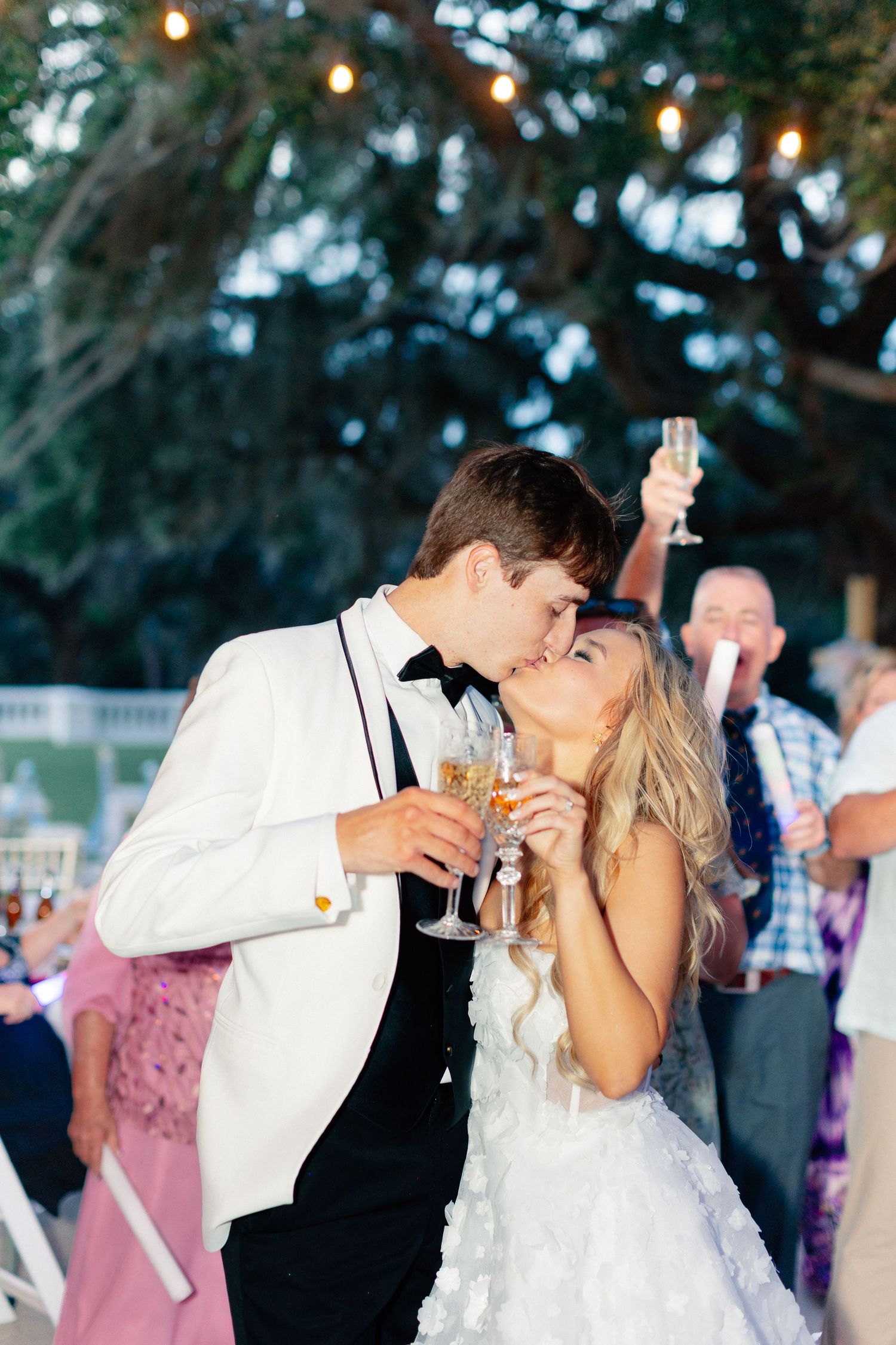 Bride and groom kissing while holding champagne glasses at outdoor wedding reception with guests celebrating in background, Florida