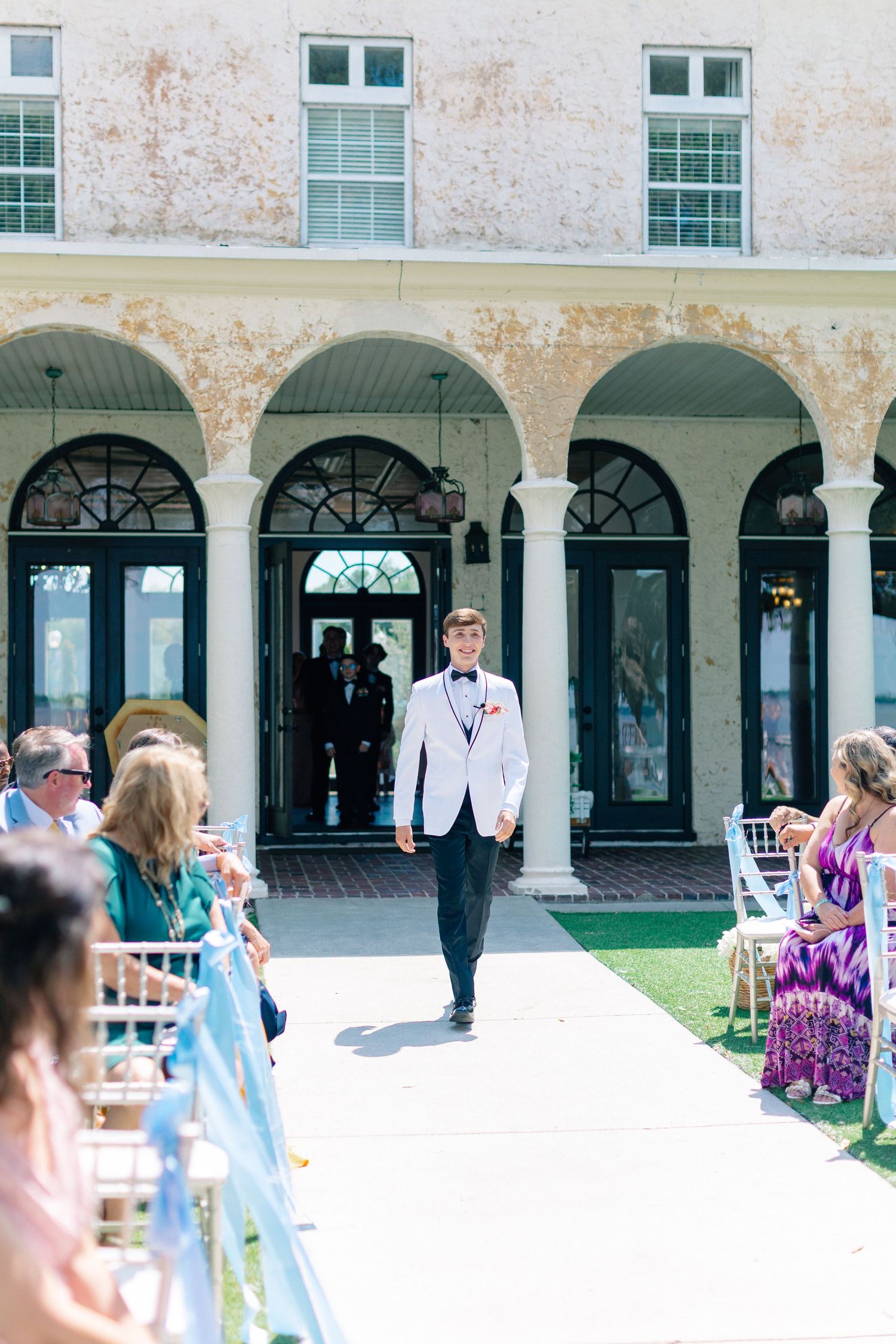 Groom in white tuxedo jacket walks down outdoor wedding aisle flanked by seated guests at elegant venue, wedding at Bella Cosa