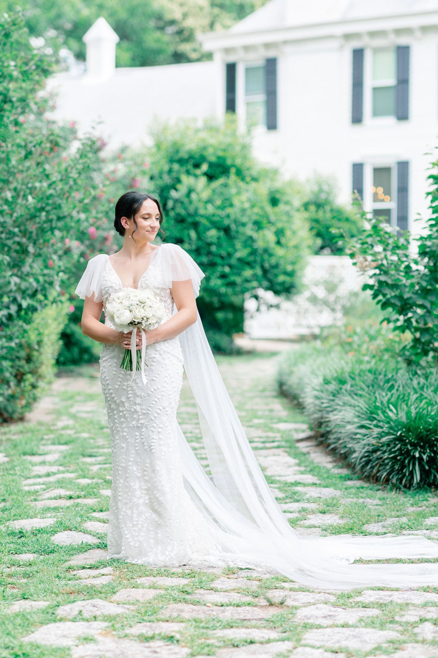Bride in beaded white gown with flutter sleeves and flowing veil holds white bouquet on stone path in lush garden.