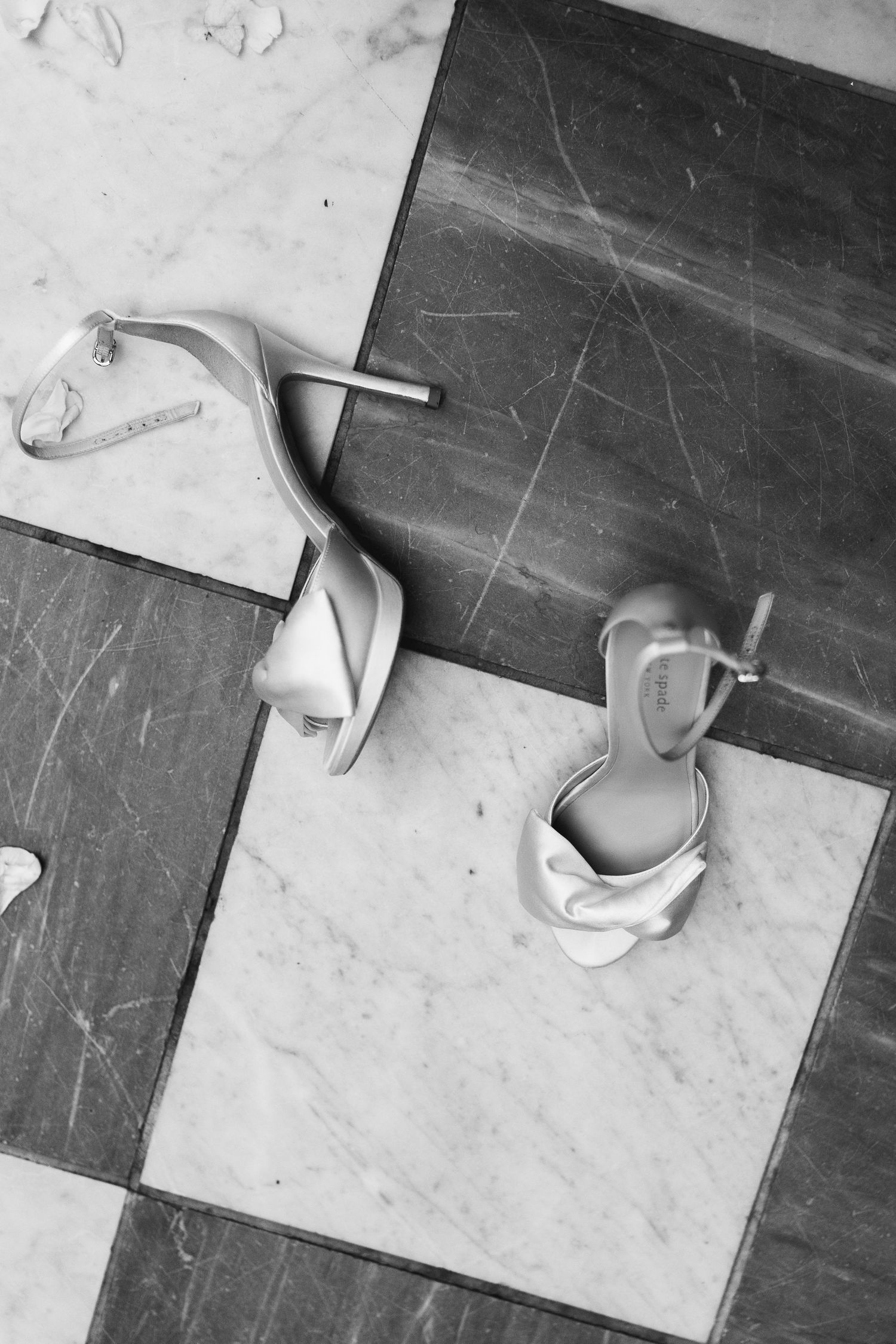 White satin strappy heels placed on a marble floor in black and white detail photography.