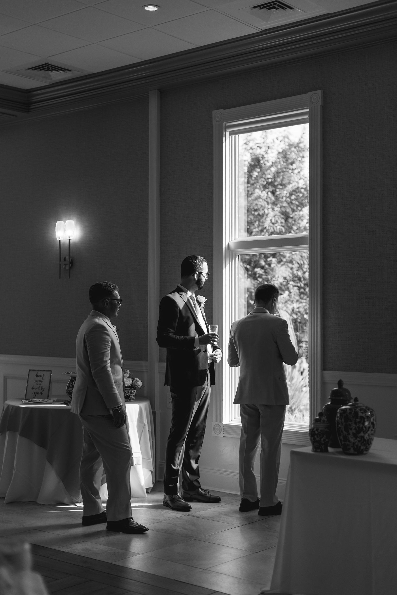 Three men in suits converse by a bright window in an elegant venue, captured in classic black and white photography.