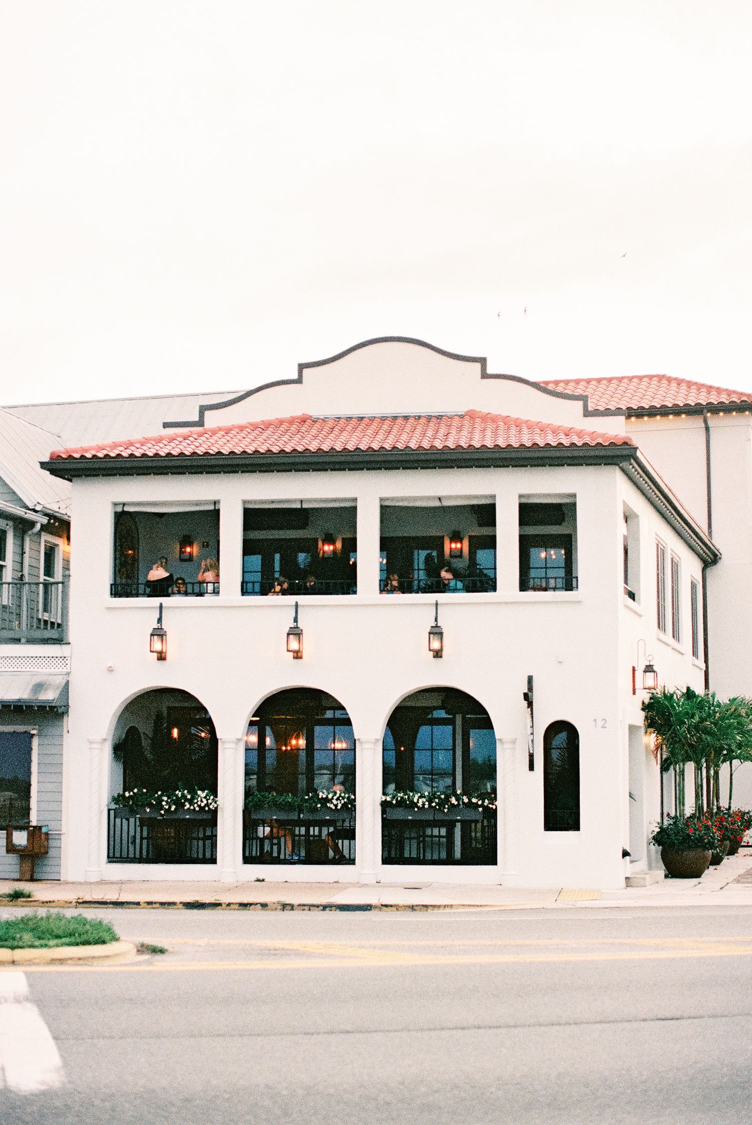 White two-story Mediterranean-style building with red tile roof, arched windows, and warm lantern lighting on a street corner.