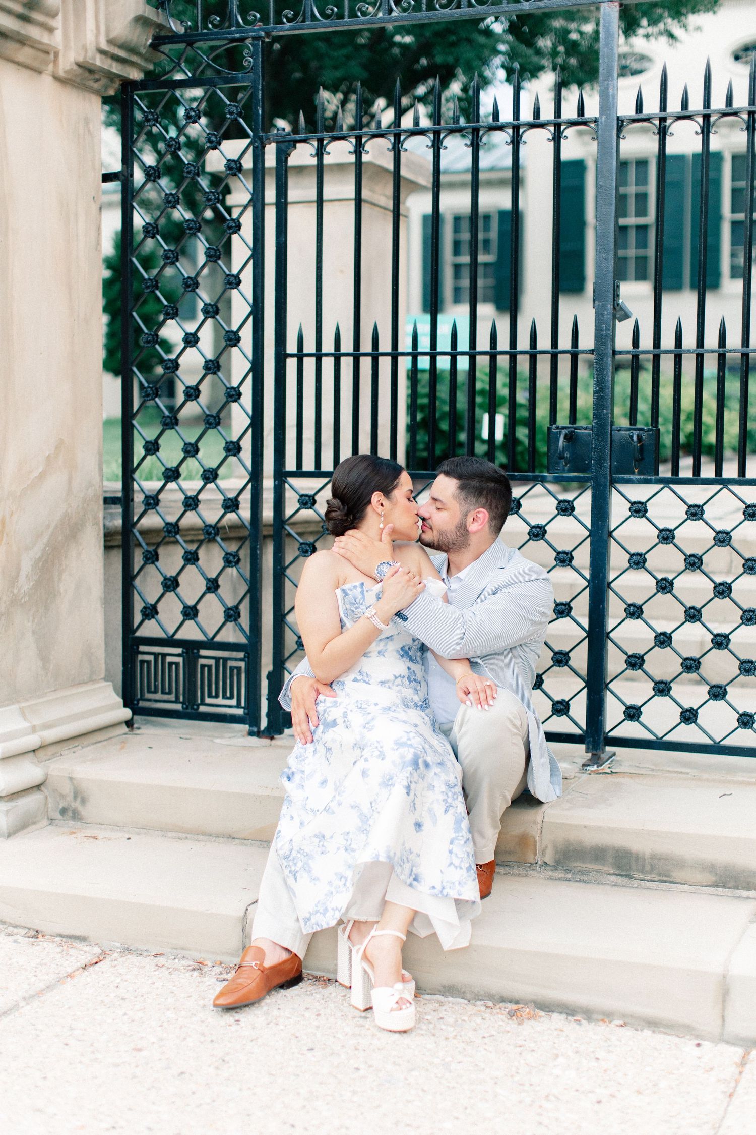 Engaged couple shares a romantic kiss while seated on steps in front of ornate black iron gates outdoors.