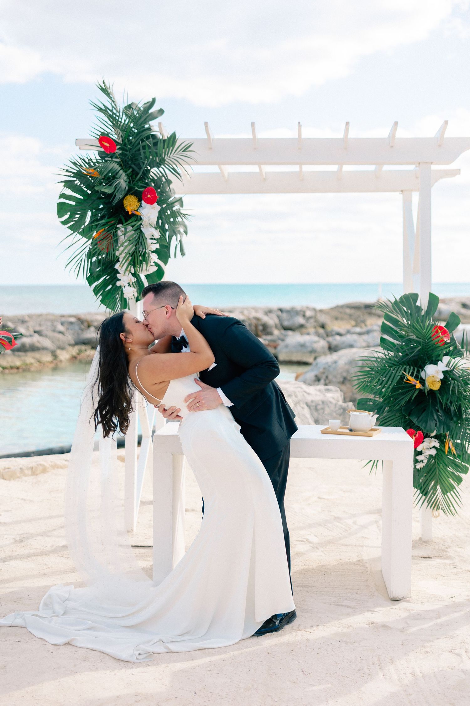 Bride and groom share a kiss at tropical beach wedding ceremony with white pergola and lush green floral arch.