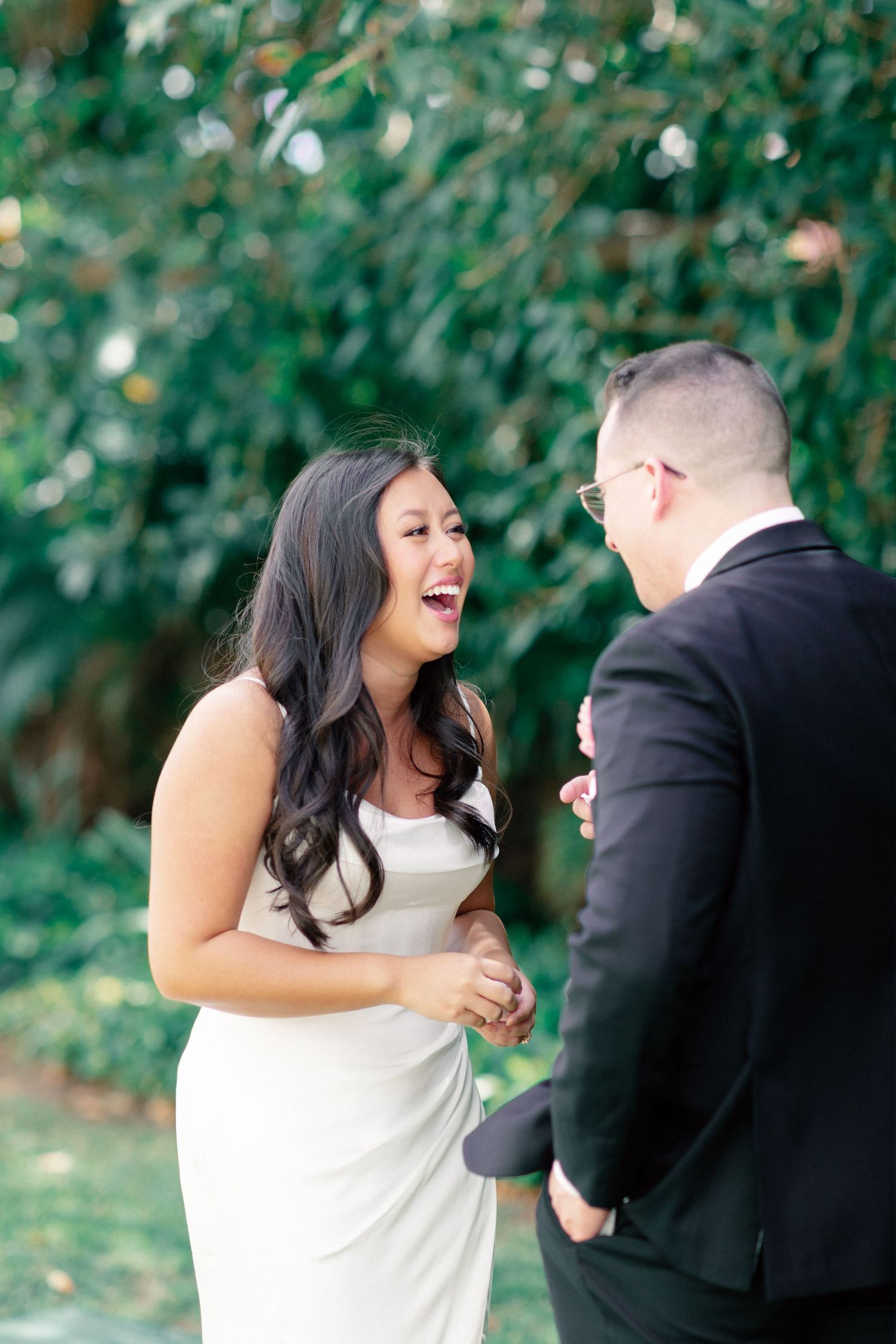 Bride in white gown laughing joyfully with groom in black suit during outdoor wedding ceremony.
