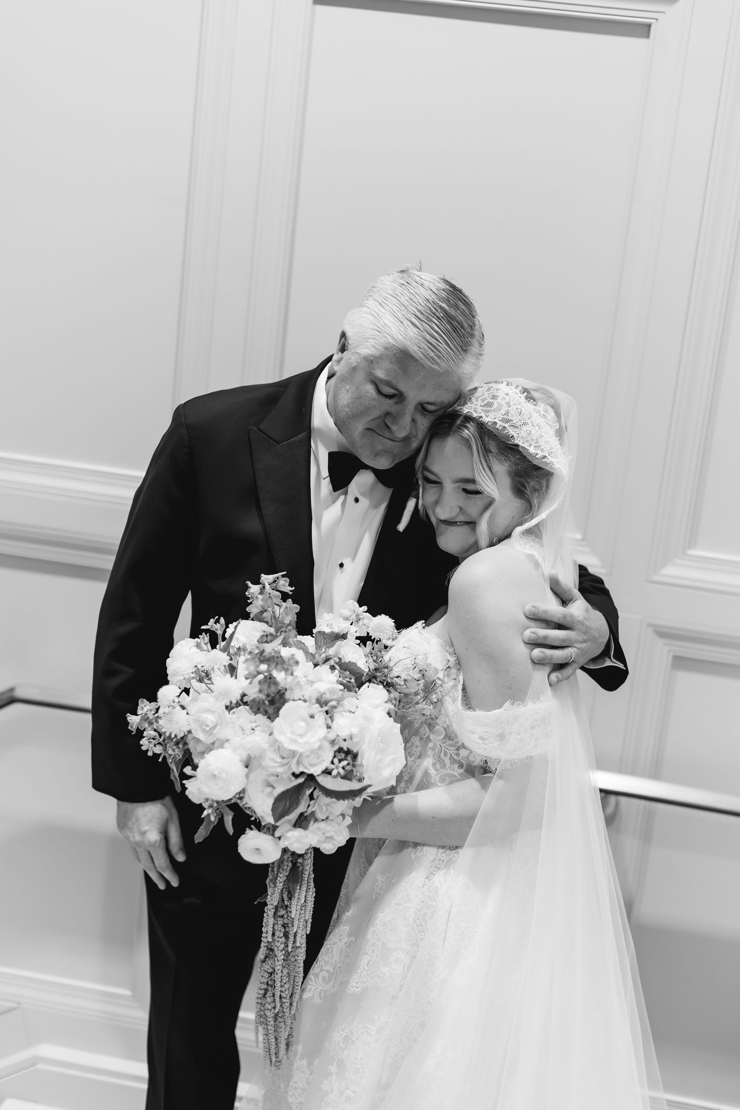 Black and white photo of a bride holding bouquet sharing tender embrace with older man in tuxedo at wedding.