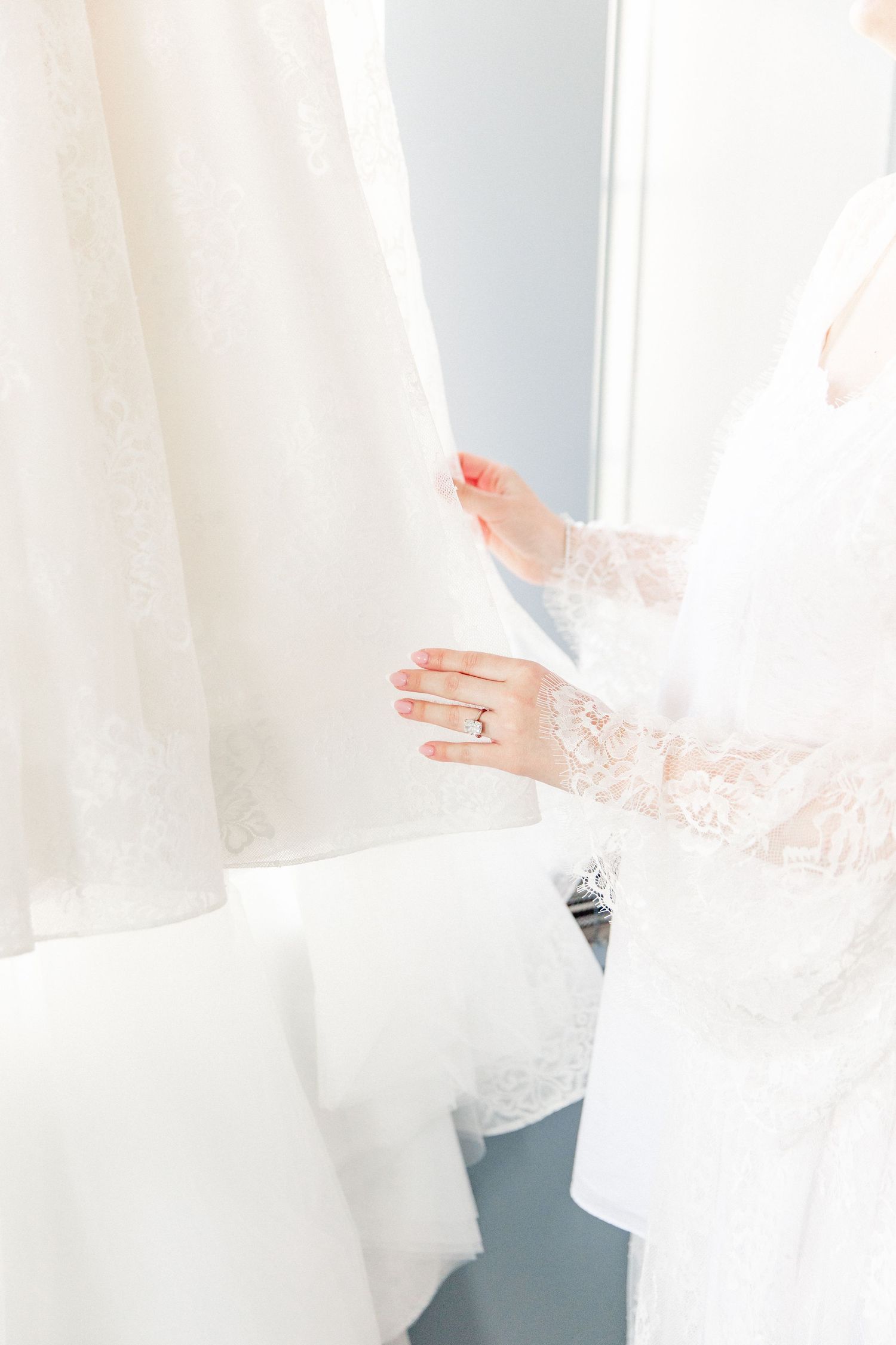 Bride in lace wedding dress admiring hanging bridal gowns, showcasing elegant diamond engagement ring on her hand.