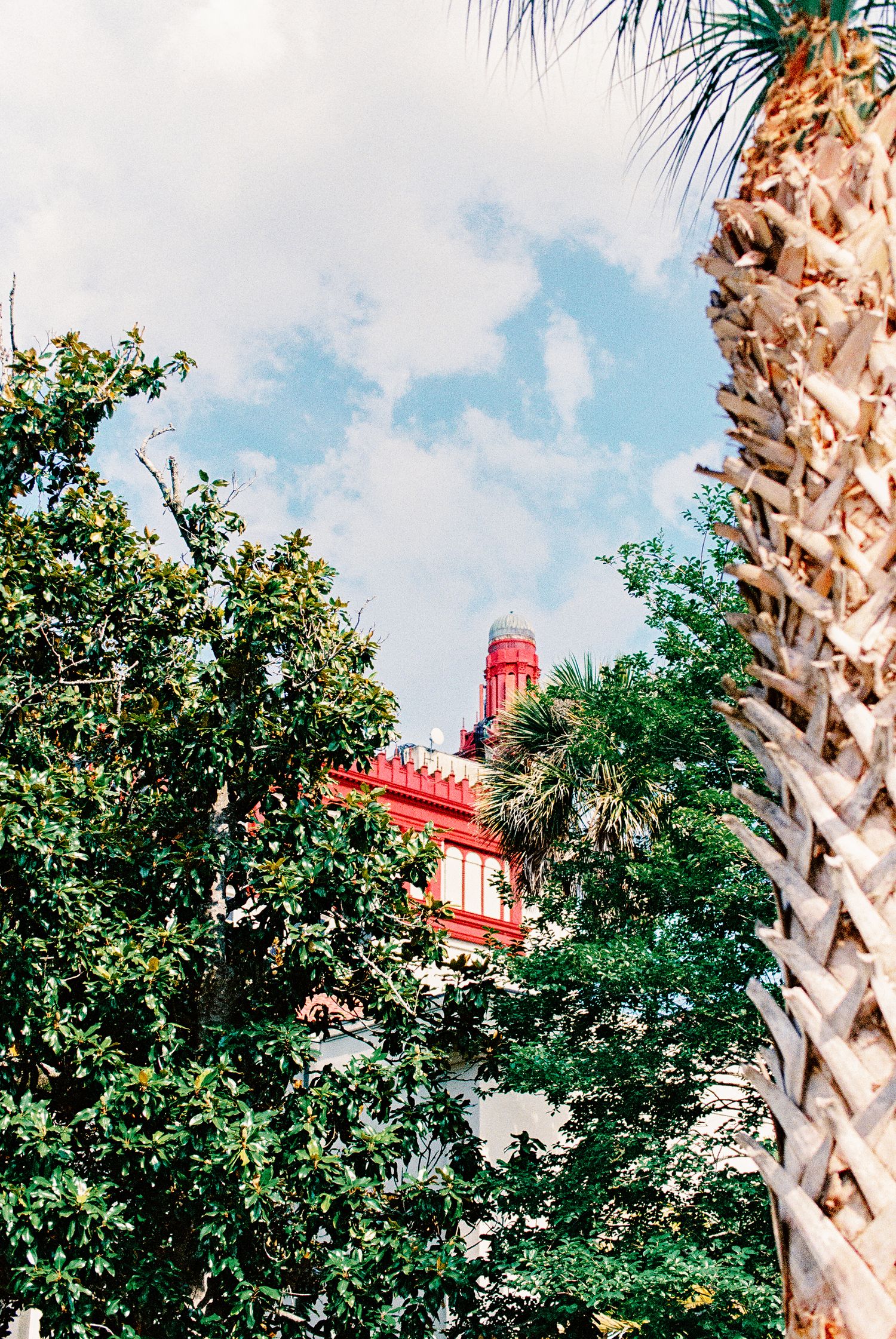 Red historic building tower peeking through lush green trees and a palm tree under a partly cloudy blue sky.