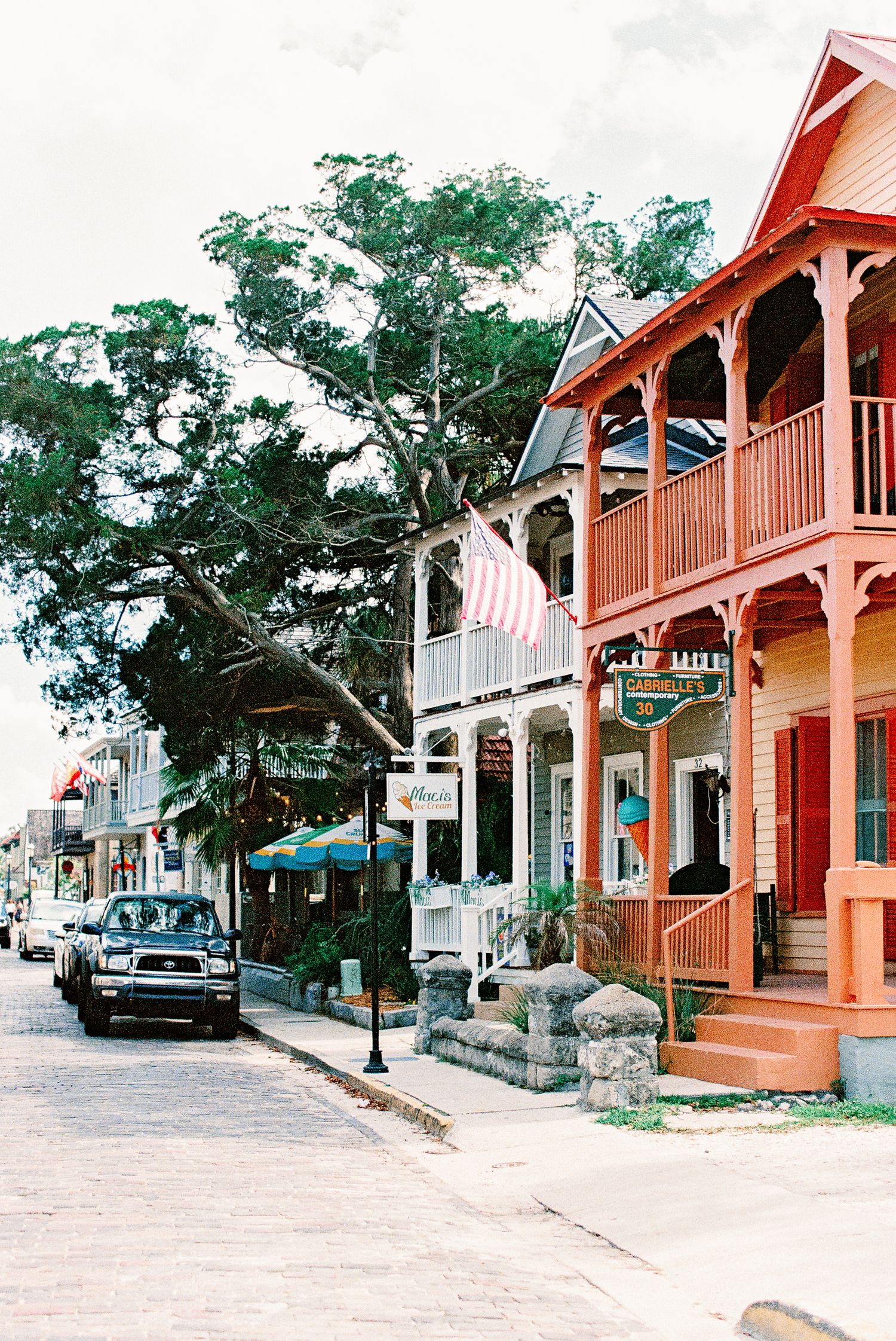 Colorful historic buildings with porches line a charming small-town street with an American flag and large oak tree.