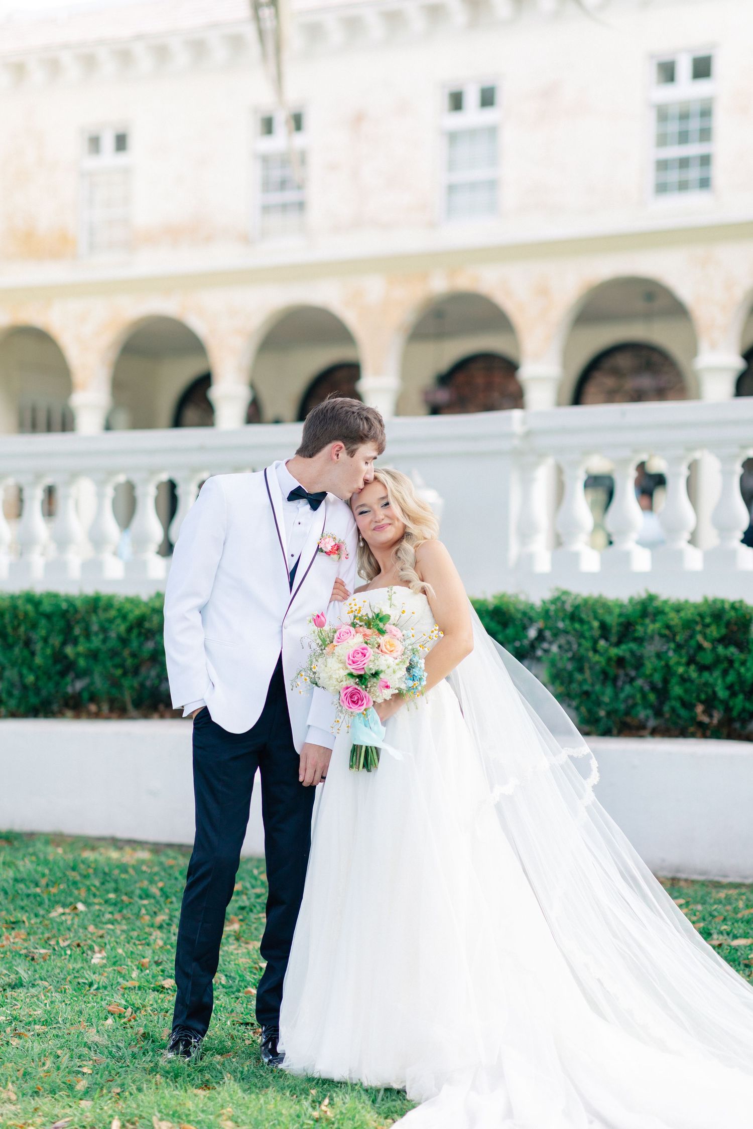 Groom in white tuxedo kisses bride in strapless ball gown holding colorful bouquet outside elegant venue, wedding at Bella Cosa