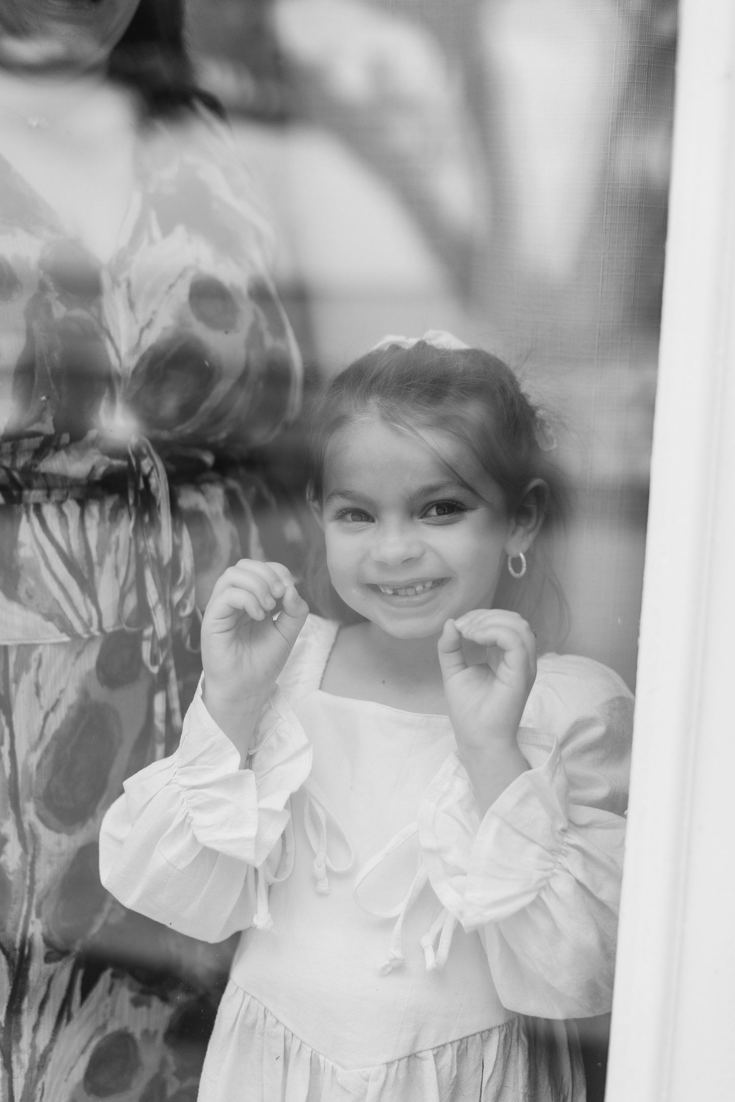 Black and white photo of a smiling little girl in a white dress with puffy sleeves, standing by a window, wedding at Bella Cosa